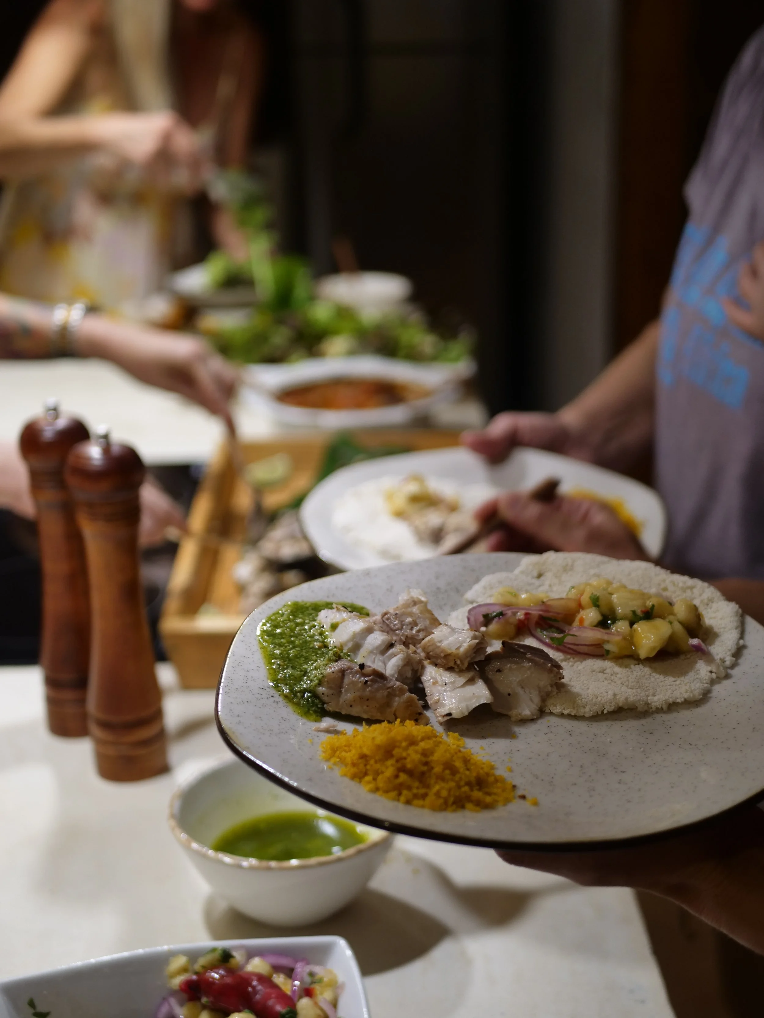 Close-up of a white plate with traditional Mexican foods: fish with green sauce, a taco with meat and vegetables, and a pile of yellow rice. In the background, there are bowls of salsa and green sauce, a mortar and pestle, and people serving themselv