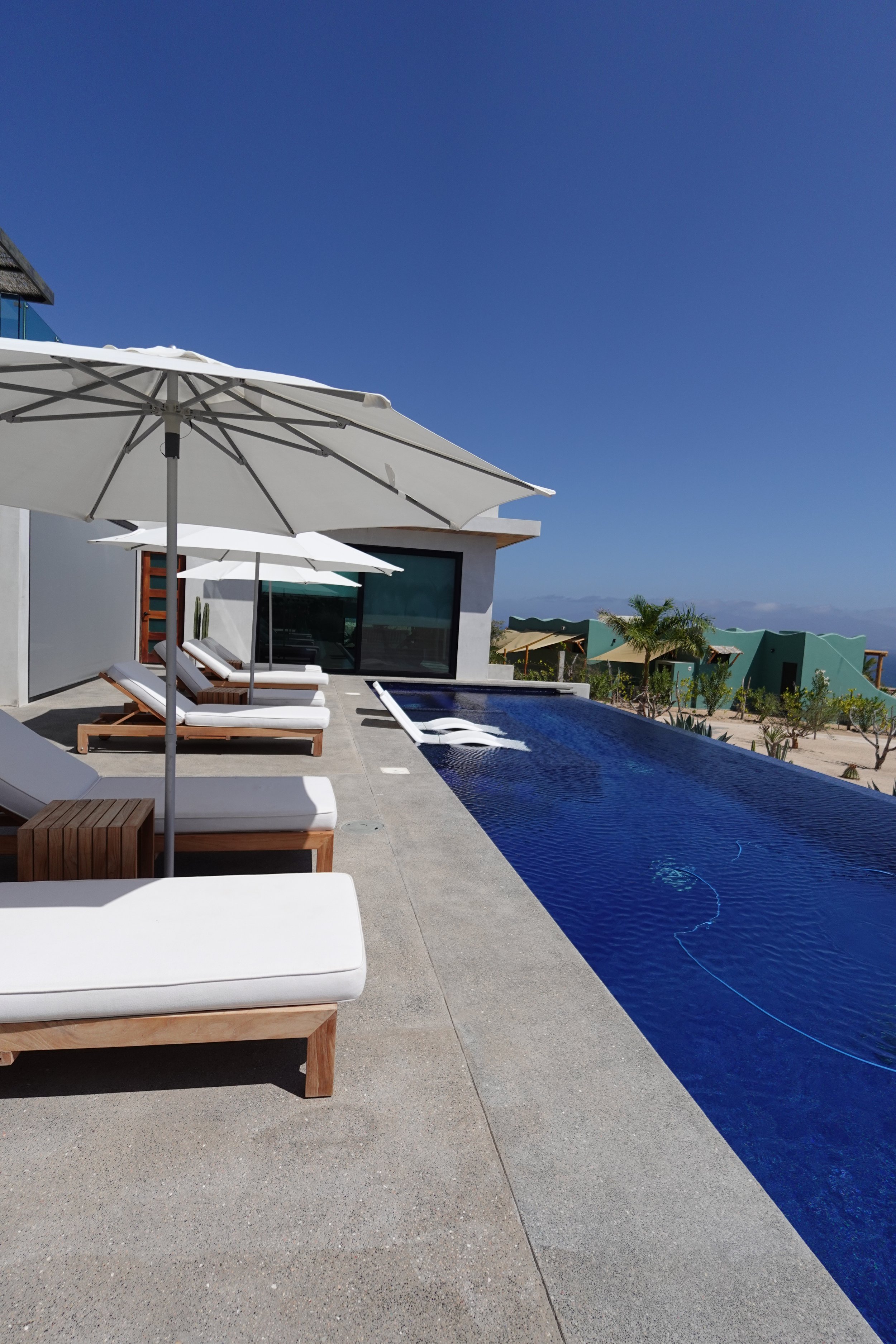 Modern outdoor poolside area with lounge chairs, large white umbrellas, a swimming pool, and a contemporary house with large glass doors under a clear blue sky.