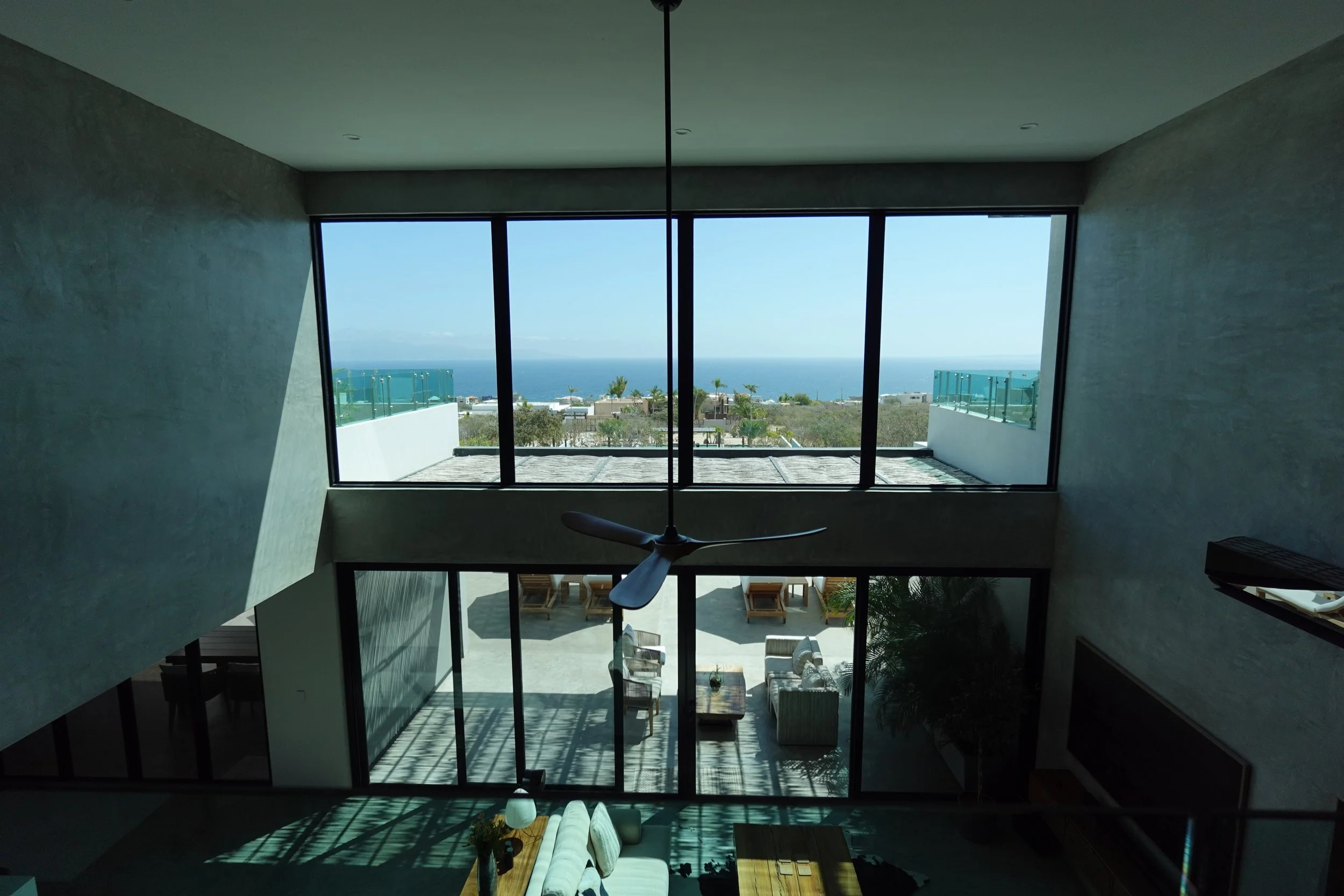 Interior of a modern house with large floor-to-ceiling windows offering a view of the ocean and coastline, with a ceiling fan hanging from the ceiling and a seating area visible on the lower level.