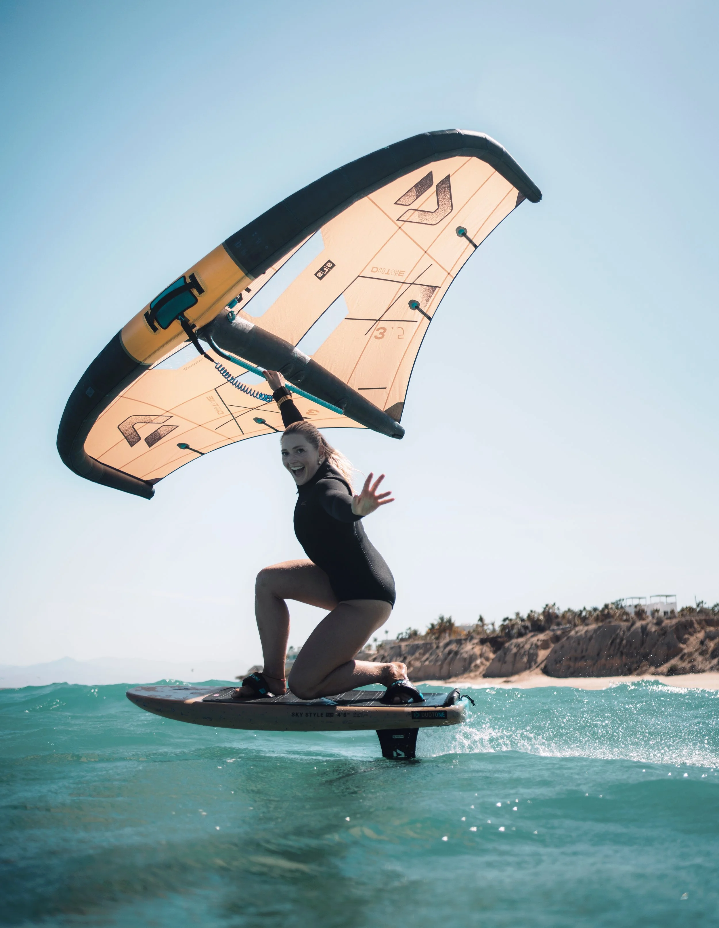 A woman with long hair, smiling and waving, stands on a stand-up paddleboard on the ocean, holding a large wing sail, with a rocky coastline and clear sky in the background.