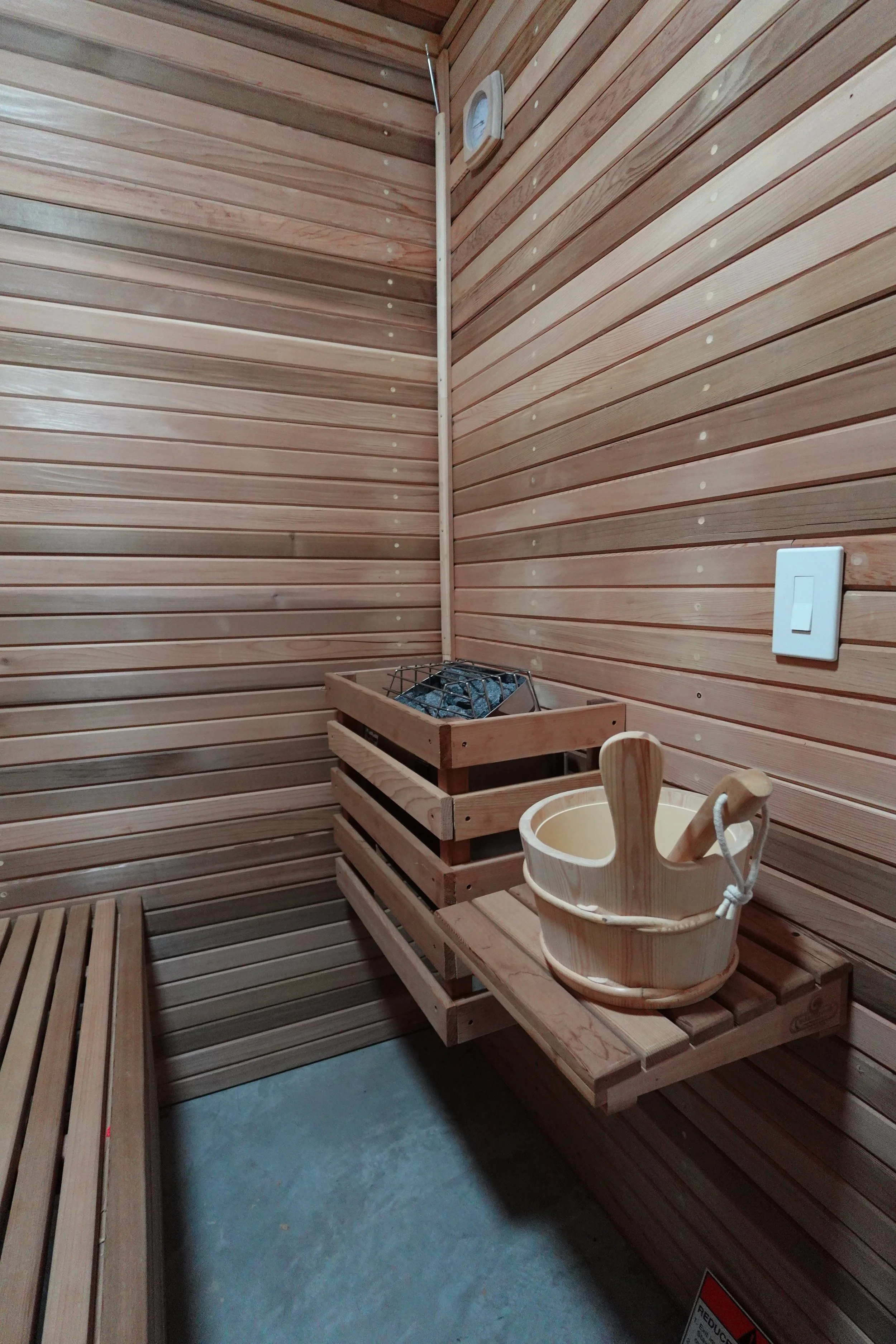 A small wooden sauna room with horizontal wooden paneling on the walls and wooden benches on the sides. There is a wooden bucket and ladle on one of the benches and a small metal heater with rocks in the corner. A white thermostat is mounted on the w