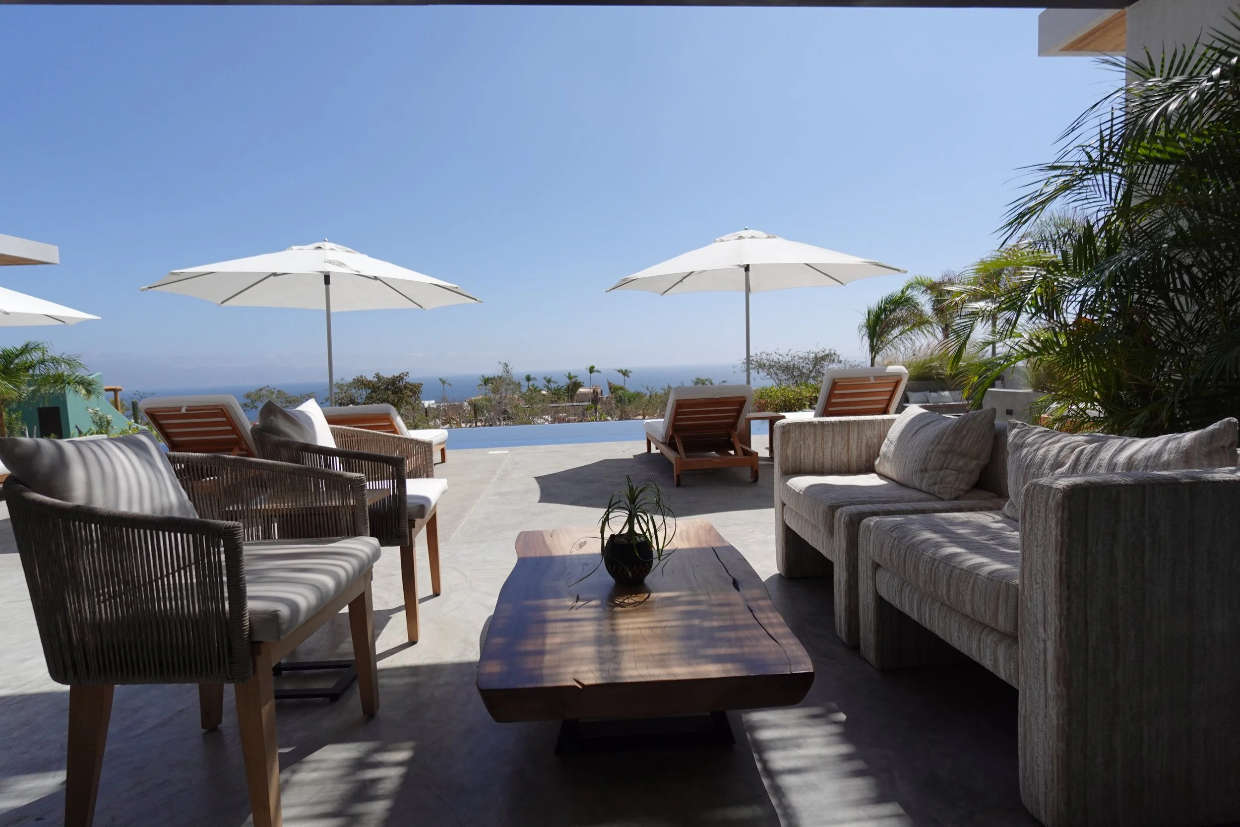Outdoor patio with wooden and cushioned furniture, white umbrellas, and a view of trees and the ocean under a clear blue sky.