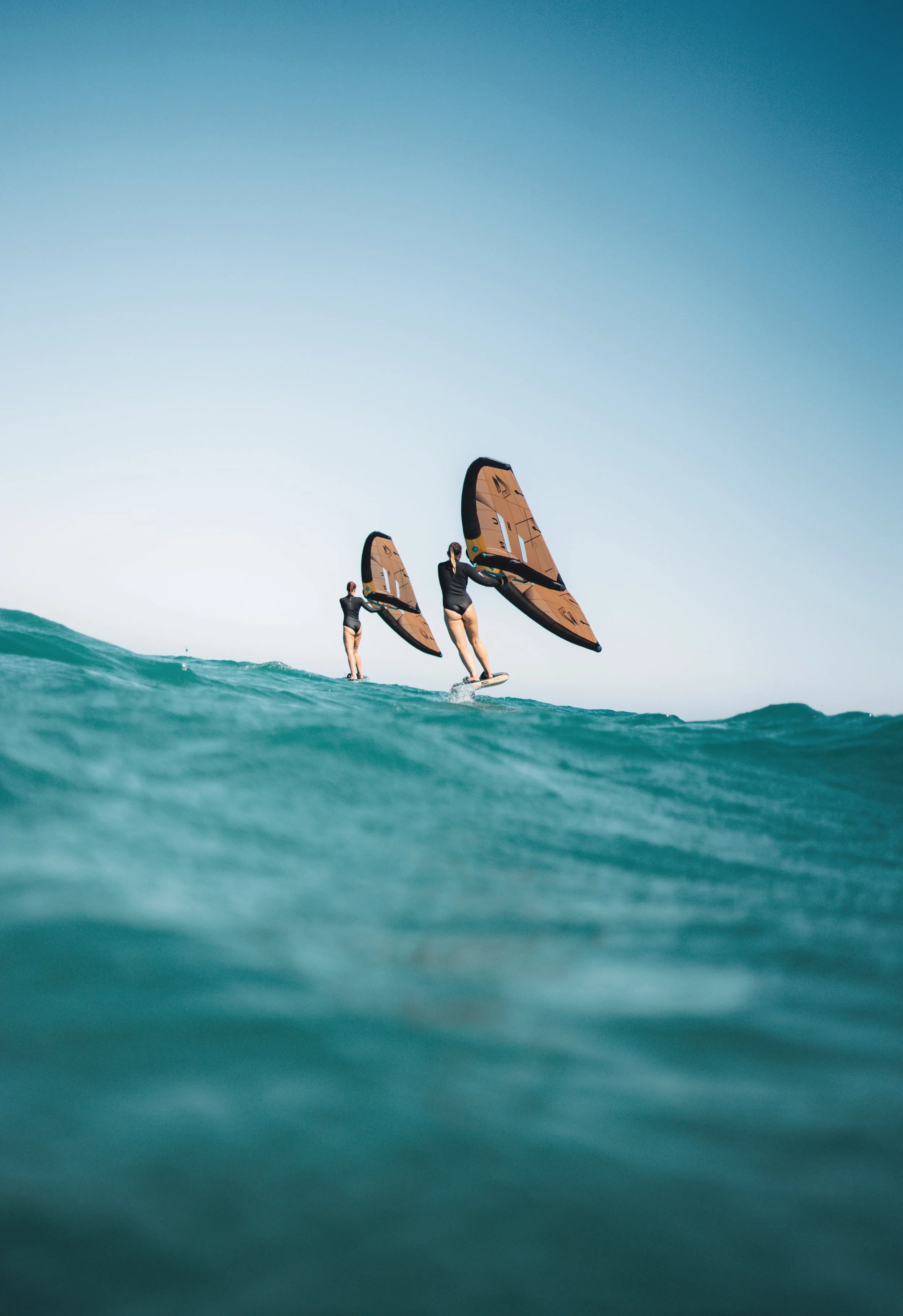 Two women are surfing on a calm ocean, holding large windsurfing sails, with clear blue sky overhead.