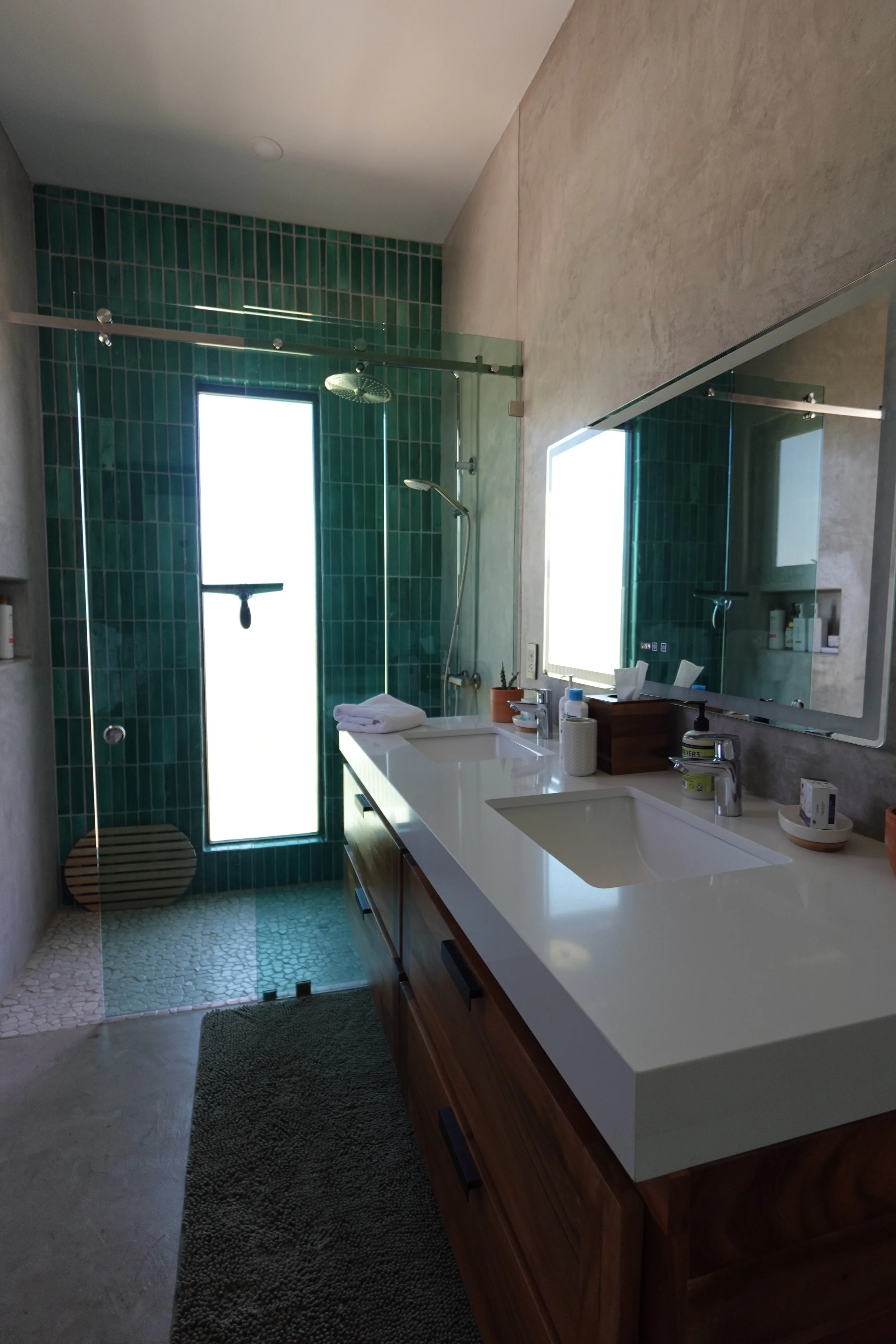 Modern bathroom with a shower area with glass door, green tiles, and a rainfall showerhead. White sink with a wooden cabinet, mirror, and various toiletries on the countertop. Towels are placed on the counter, and a small gray rug on the floor.