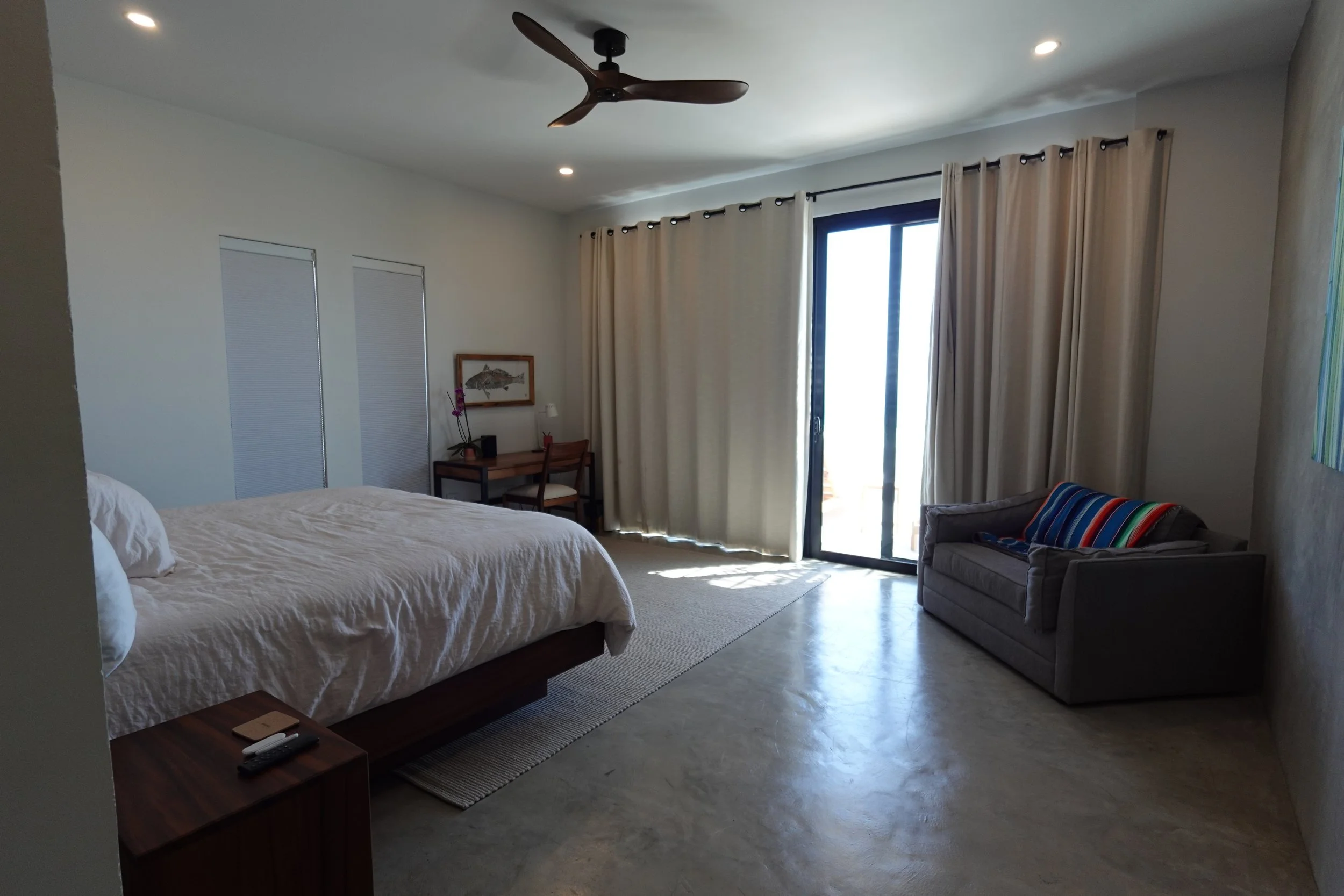 A bedroom with a large bed on the left, a small wooden desk and chair with a framed fish picture above it, large sliding glass door with beige curtains, a gray armchair with colorful striped blanket, and a ceiling fan.