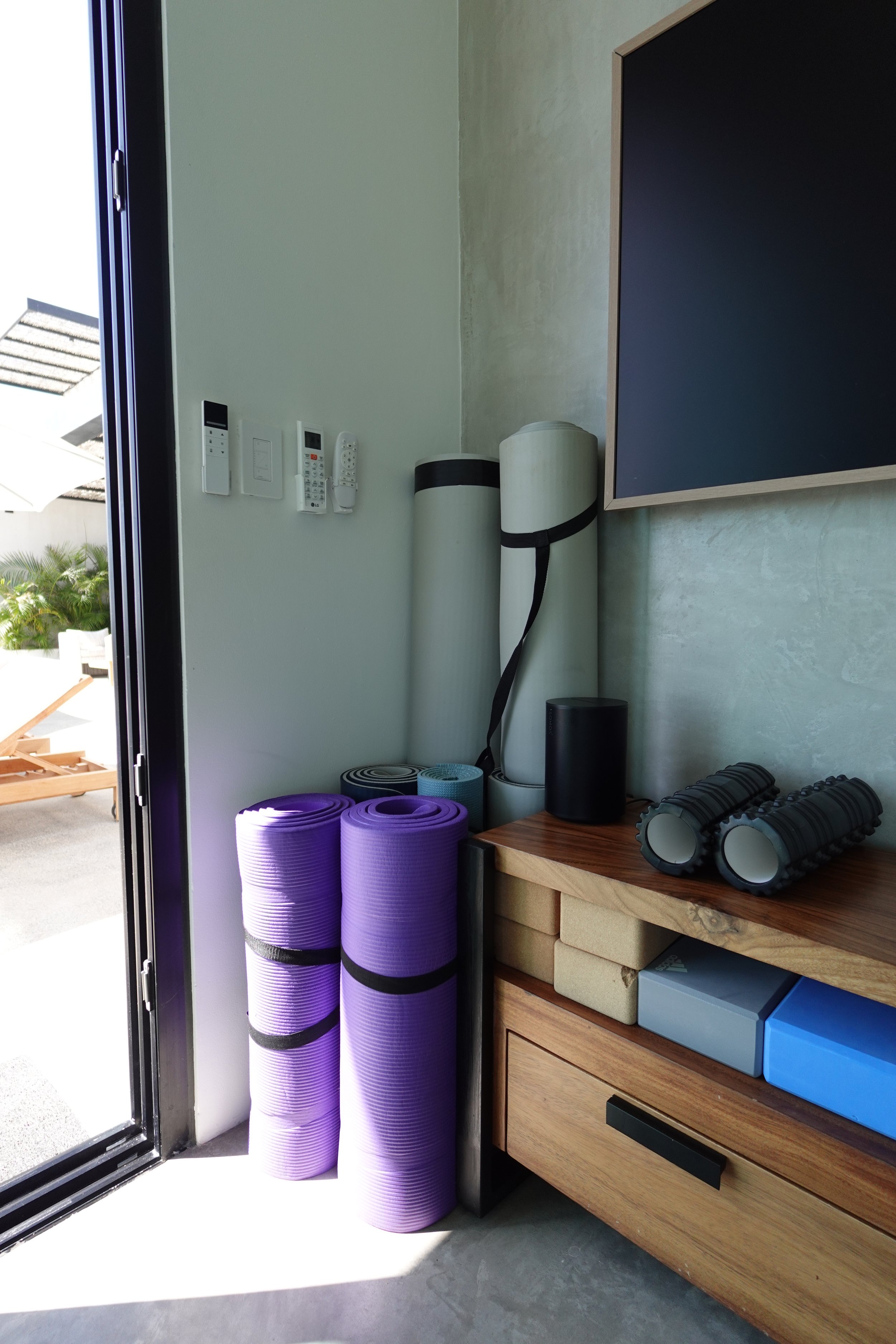 Purple yoga mats rolled up and stacked next to a wooden storage unit in a fitness room, with part of the open door and outside view visible.