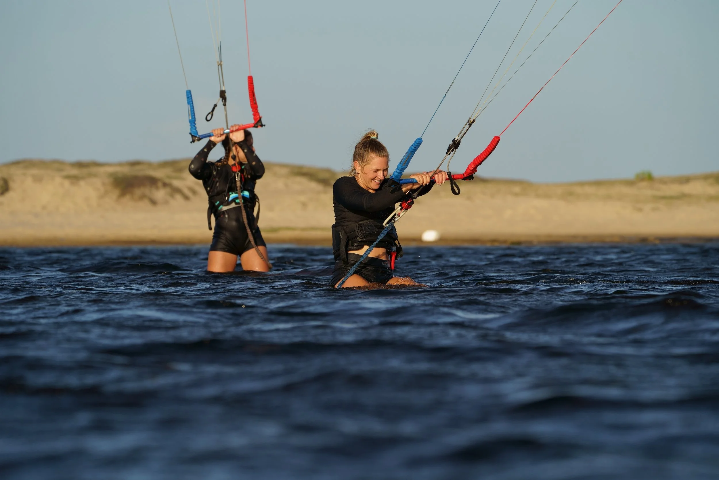 Two women in black wetsuits flying kites over a body of water during sunset.