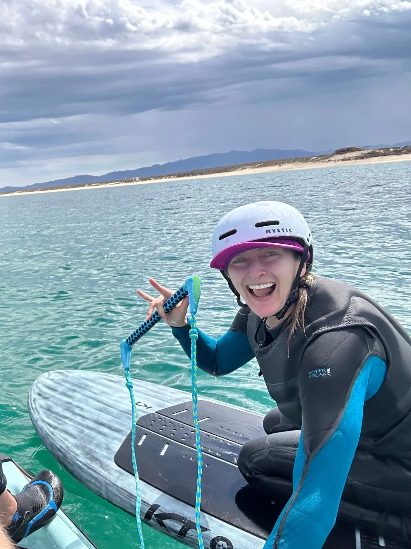 Woman in wetsuit and helmet smiling on a paddleboard in water, holding a paddle and making a peace sign, with a cloudy sky and distant land in the background.