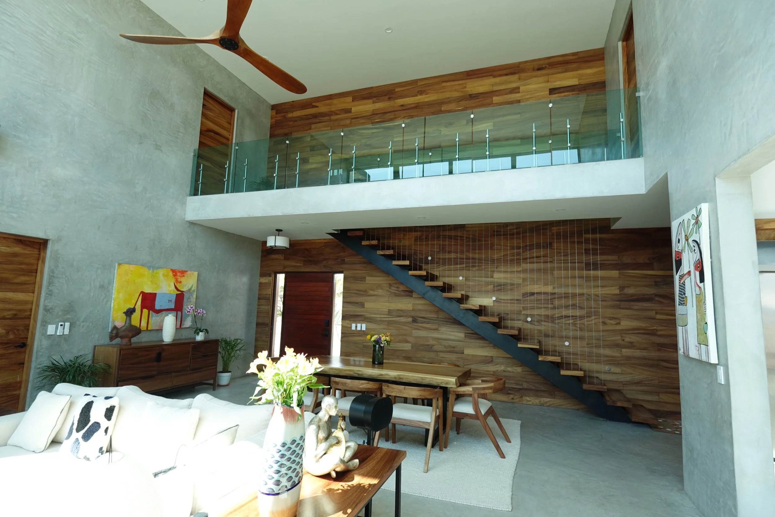 Interior view of a modern living space with a staircase, wooden accents, glass railing, ceiling fan, artwork, and various decorative items.