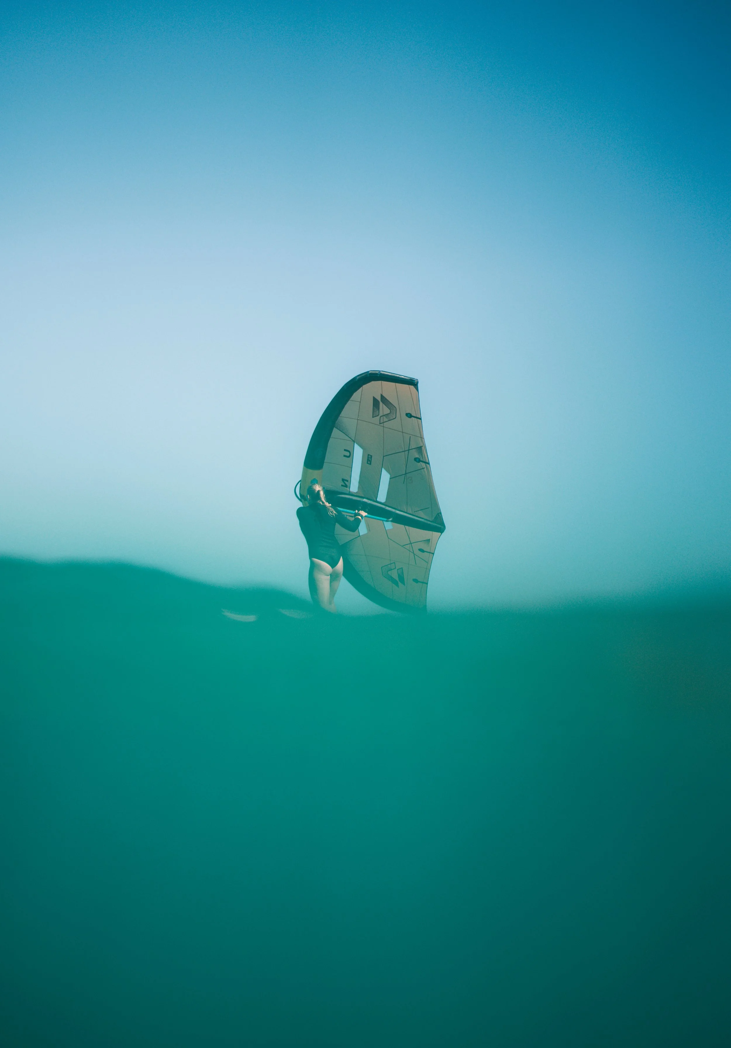 A person standing in the water holding a sailboard with a sail, under a blue sky.