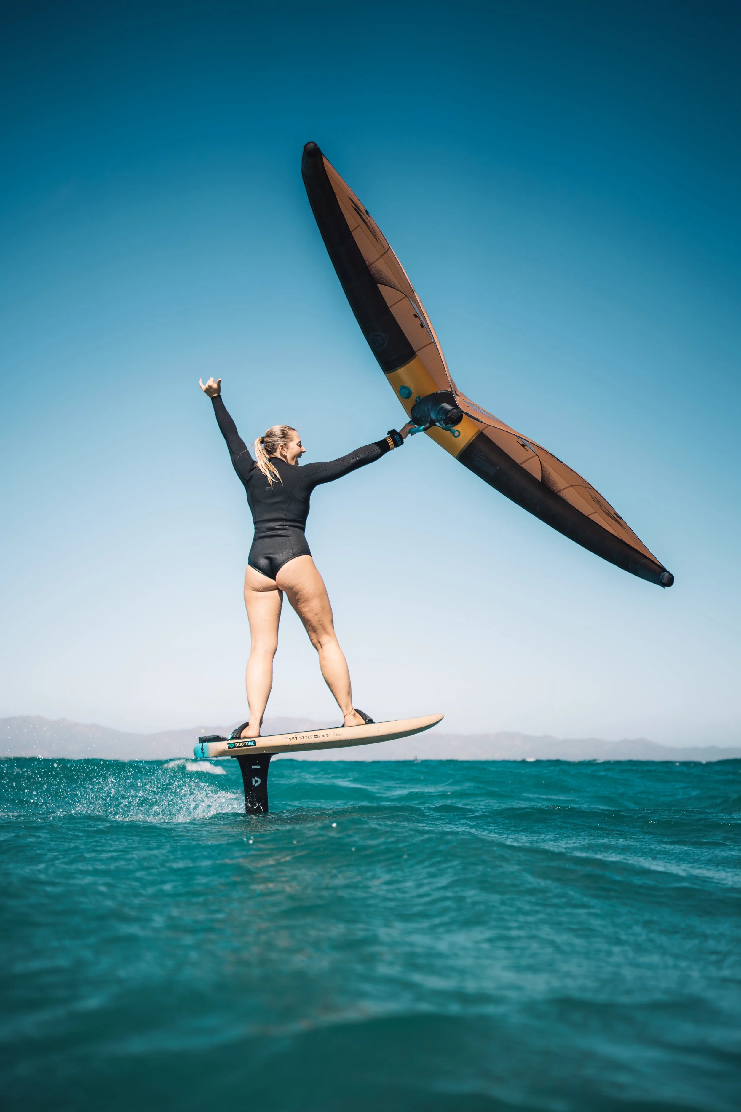A woman water surfing on a hydrofoil board, holding a large paddle, as she turns her head to the side against a clear blue sky.