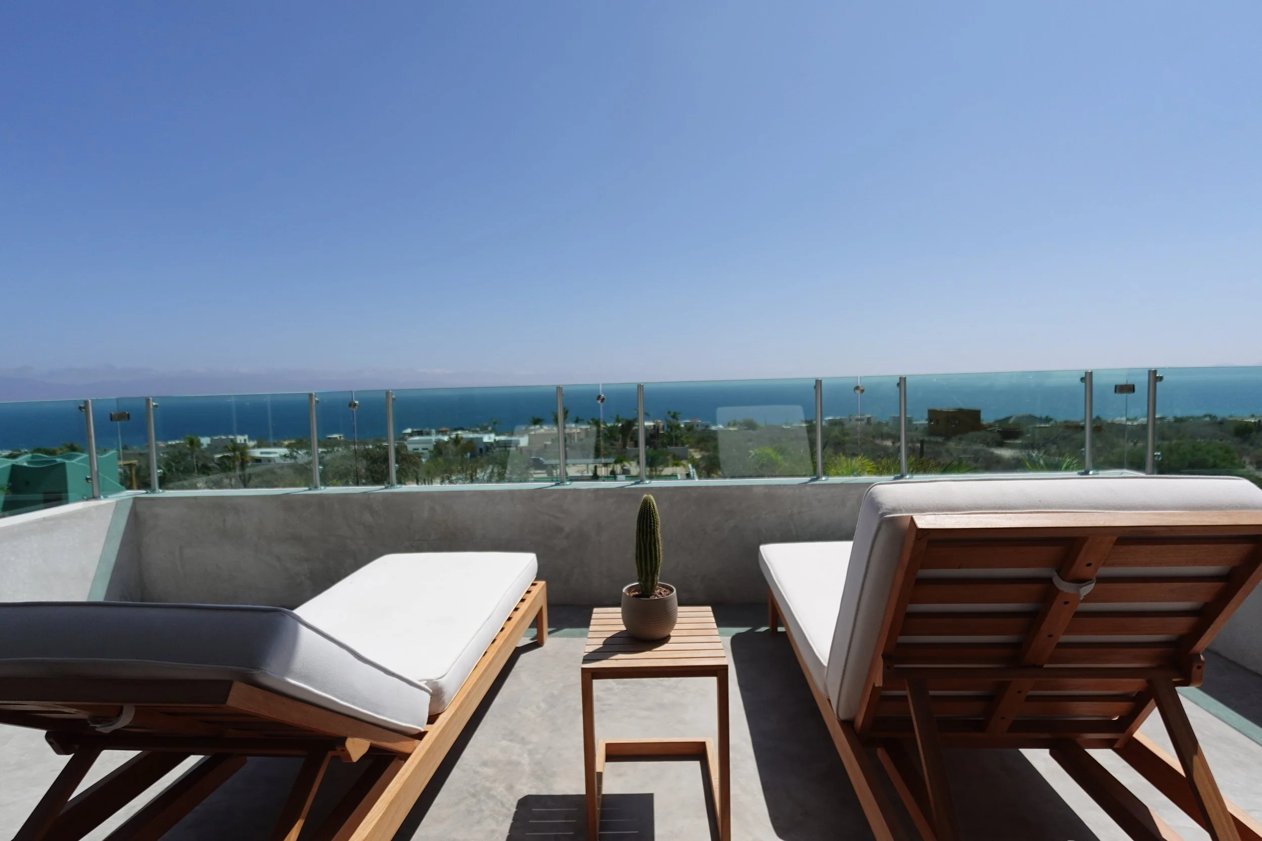 Balcony with two lounge chairs, a small wooden table with a cactus plant, glass railing, ocean view, blue sky, and distant mountains.