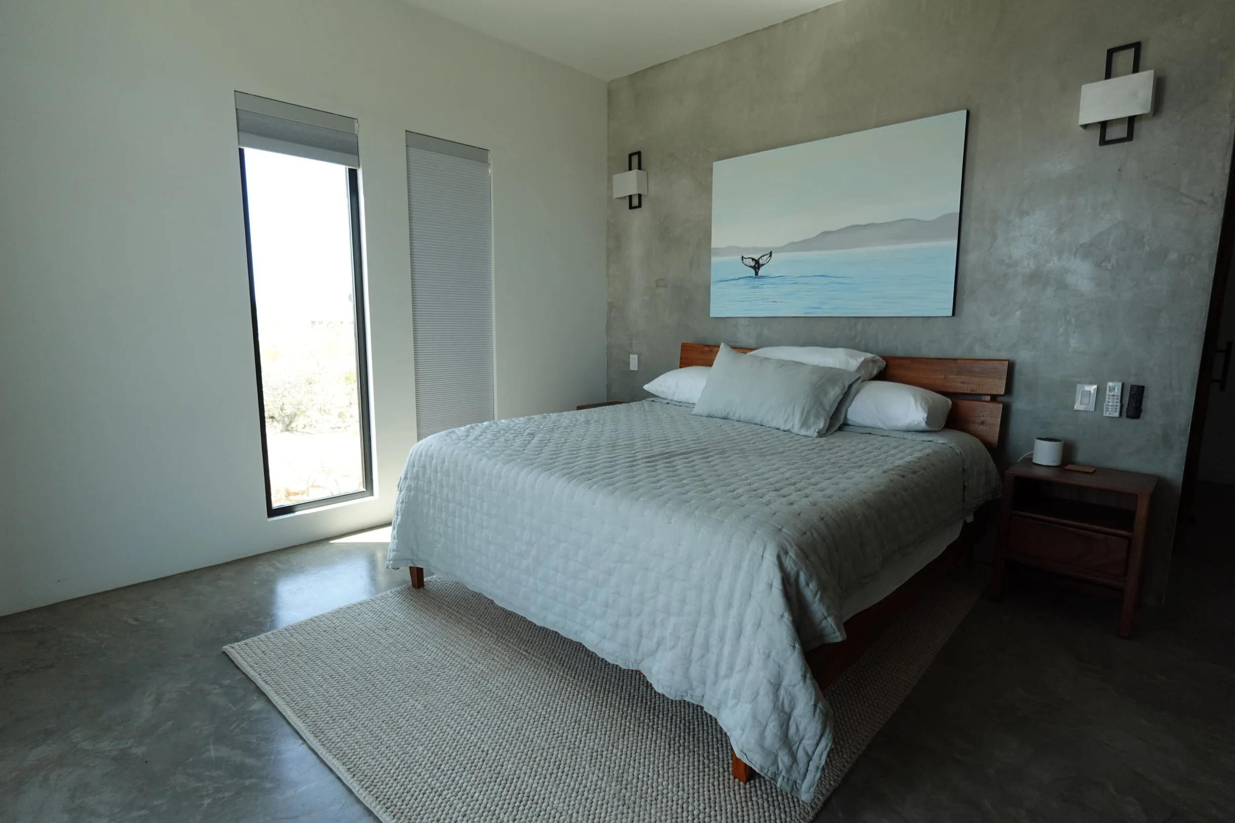 Modern bedroom with a large bed, white quilt, and pillows, featuring a concrete accent wall with a whale painting, next to a window with blinds, and a small wooden nightstand with electronic devices.
