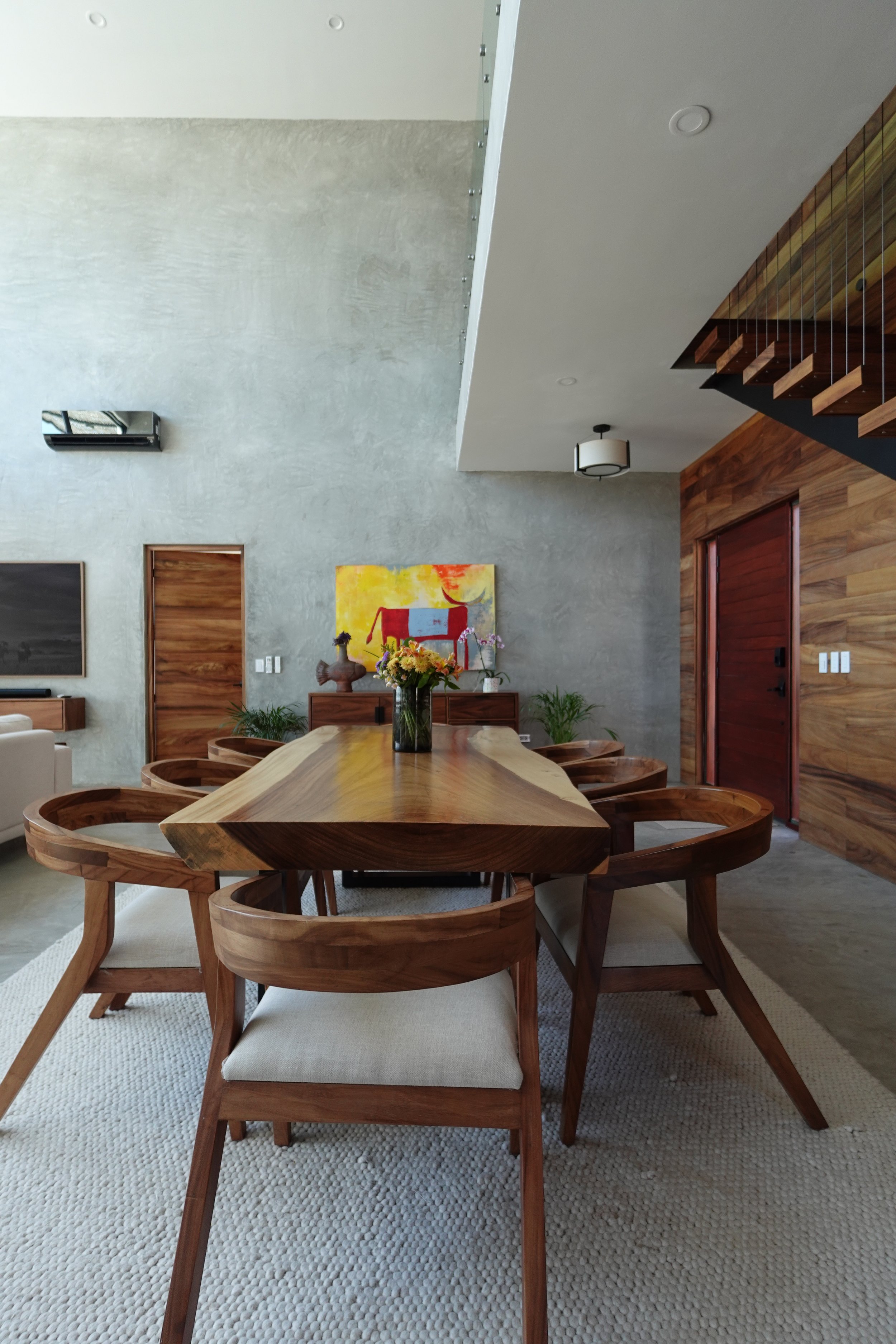 A modern dining room with a wooden dining table surrounded by matching wooden chairs with beige cushions. The room has a concrete wall, a textured white rug, and wooden accents on the walls and staircase. There are decorative flowers on the table, ar