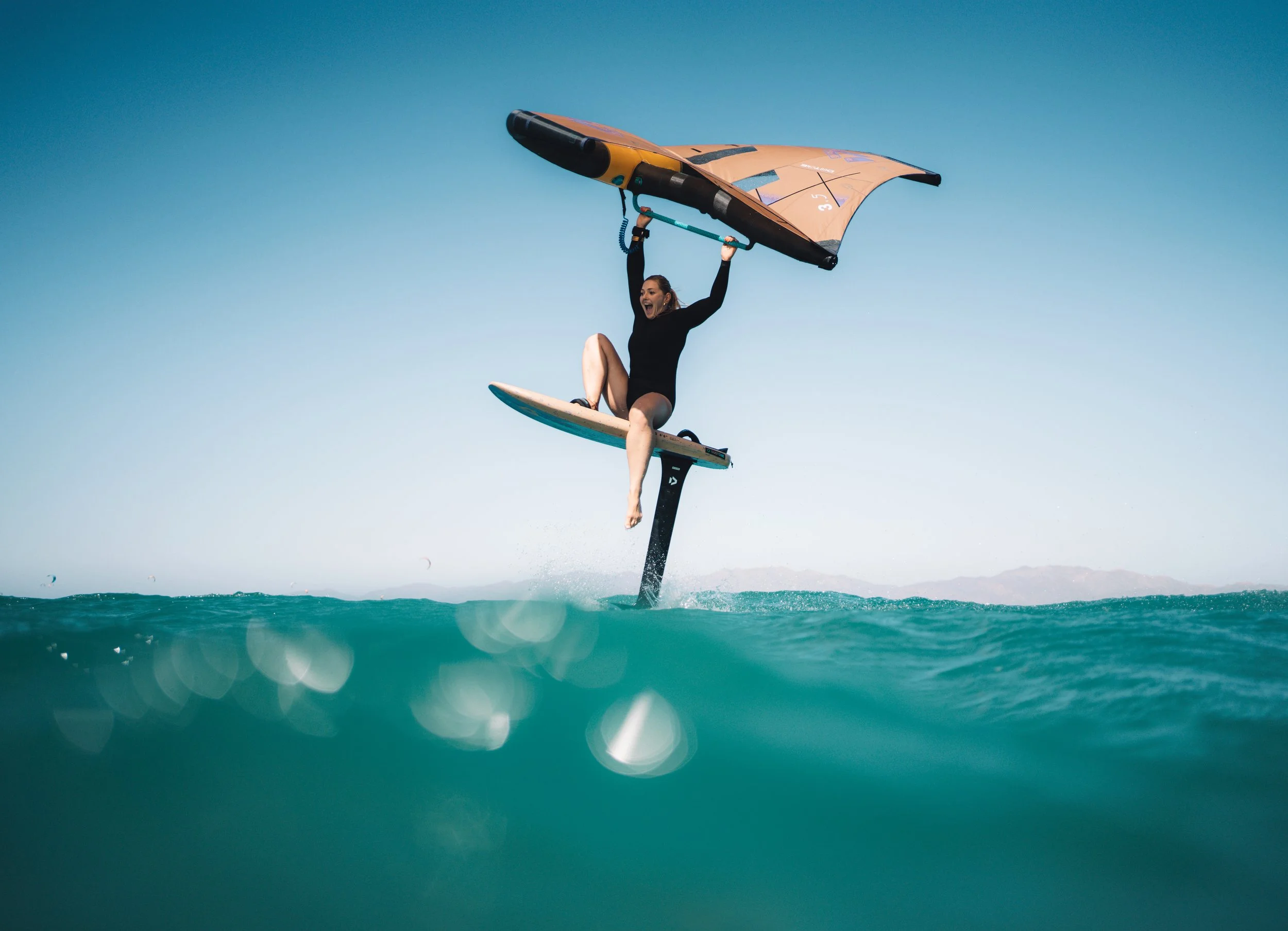 A woman wearing a black wetsuit windsurfing on a calm ocean, holding onto the sail's boom with her arms raised and smiling, with a clear blue sky above.