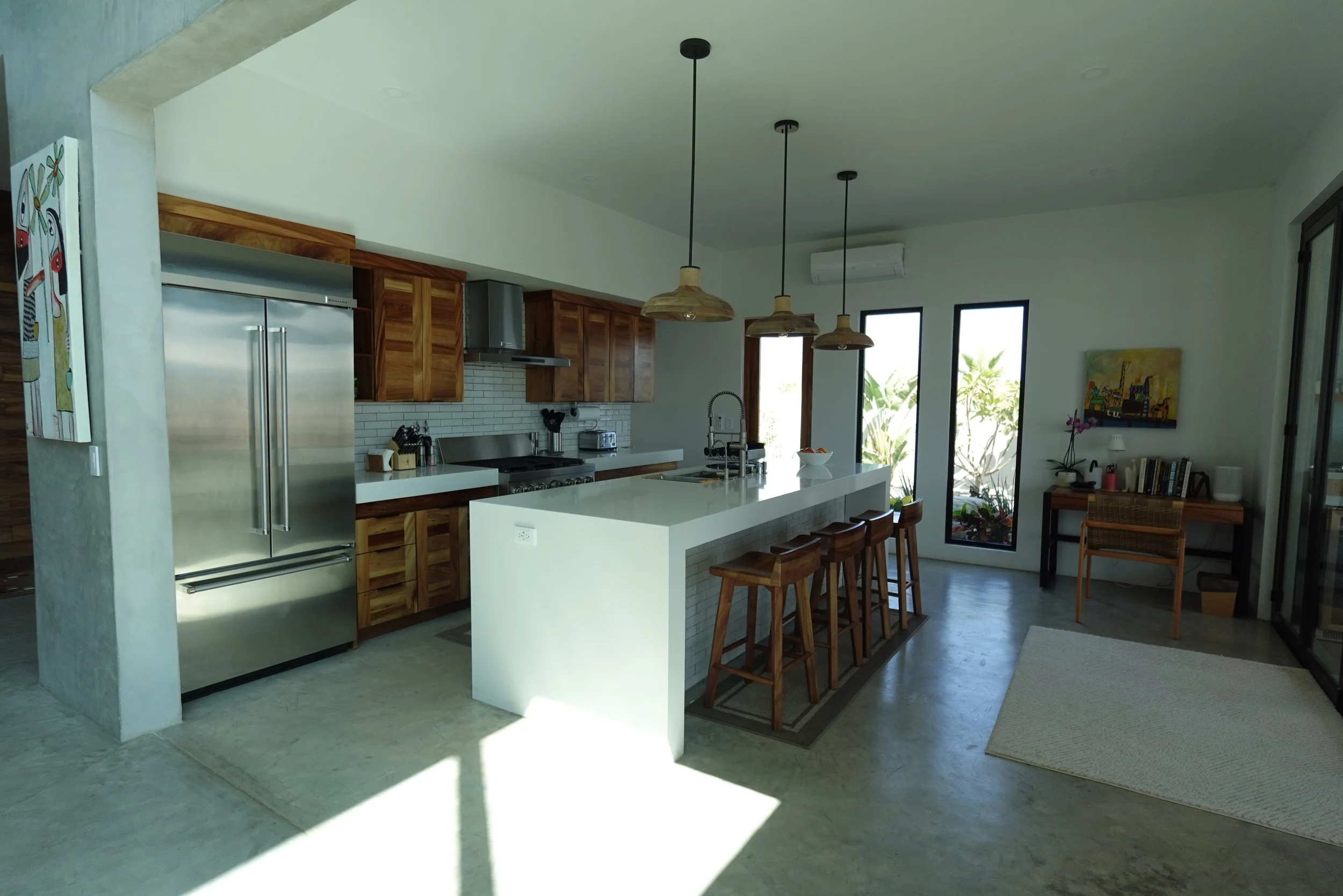 Modern kitchen with white island, wooden cabinetry, stainless steel appliances, pendant lights, large windows, and minimal decor.