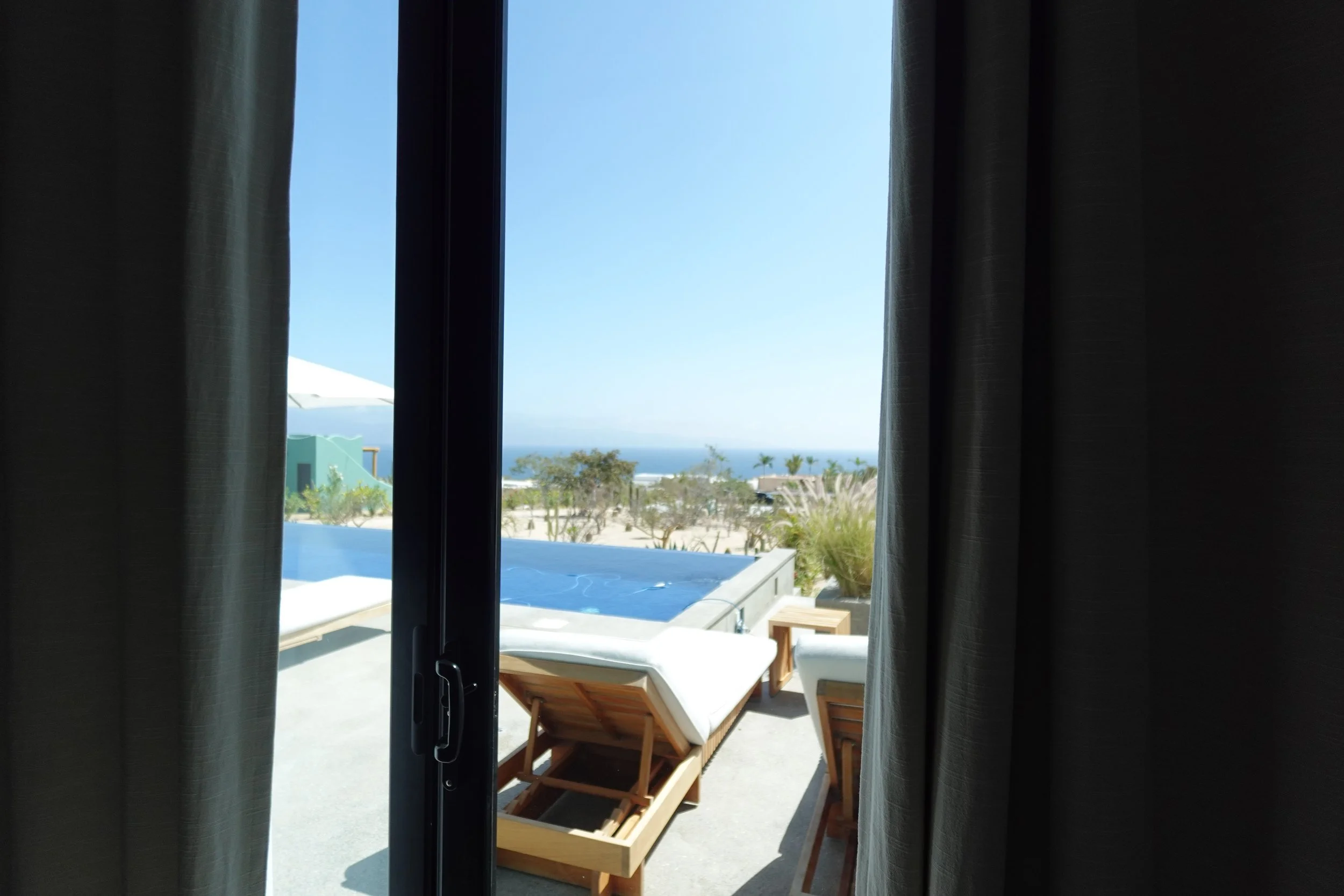 View through a sliding door shows an outdoor patio with two lounge chairs, a swimming pool, palm trees, and a clear blue sky.