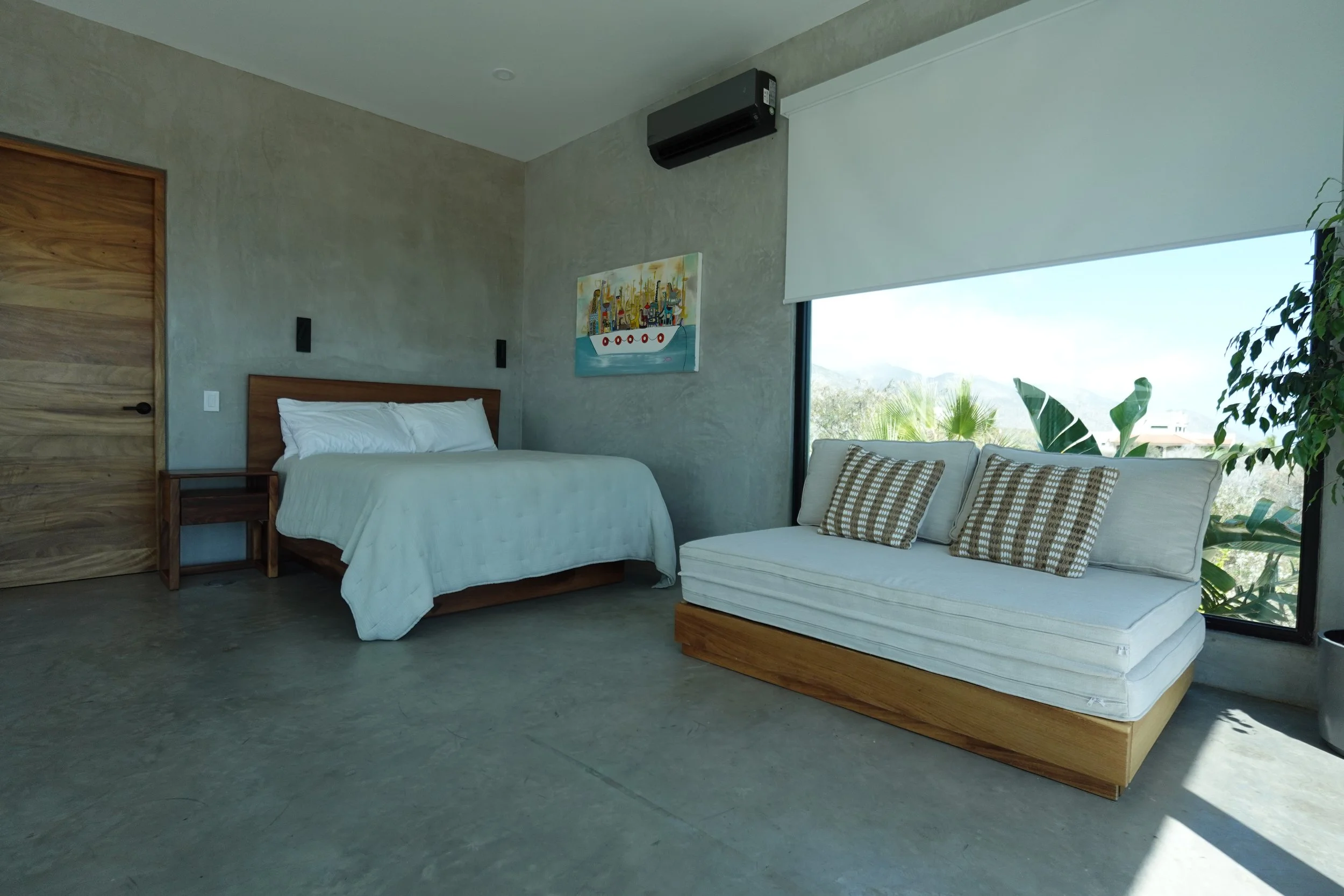Modern bedroom with a bed, a sofa, and large windows showing outdoor greenery.