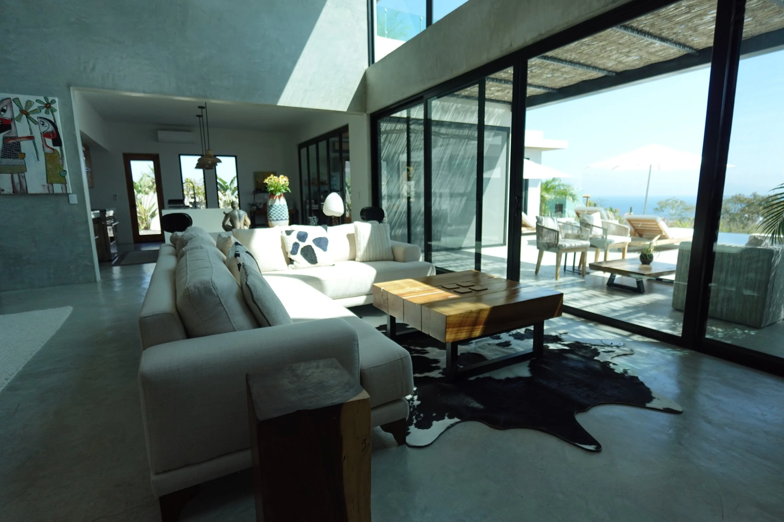 Modern living room with large glass sliding doors that lead to an outdoor patio with sun loungers and umbrellas, and an ocean view in the background.