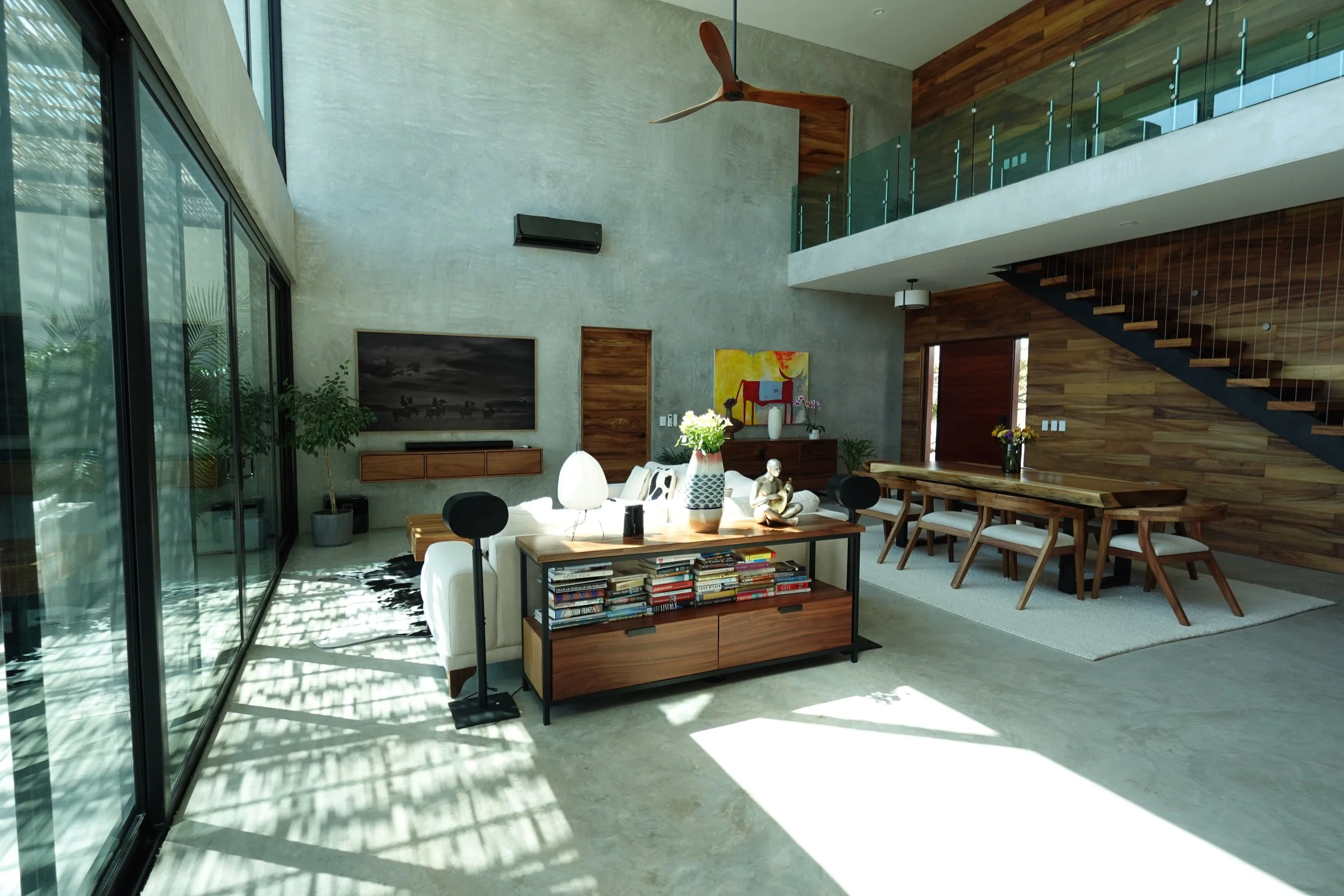 Modern open-concept living room and dining area with large glass sliding doors, concrete and wooden wall accents, a ceiling fan, a mounted television, artwork, plants, and furniture including a sofa, coffee table, and dining table with chairs.