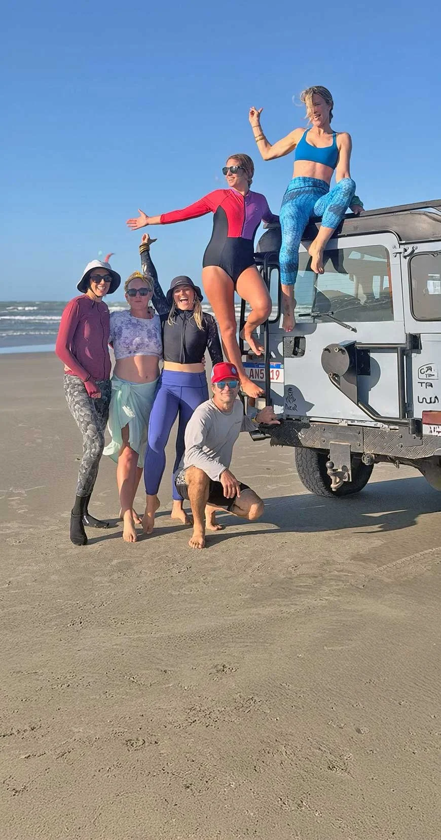 Group of people on the beach gathered around a vehicle with some sitting on it, others standing or crouching on the sand, with the ocean in the background and clear blue sky, enjoying a sunny day.
