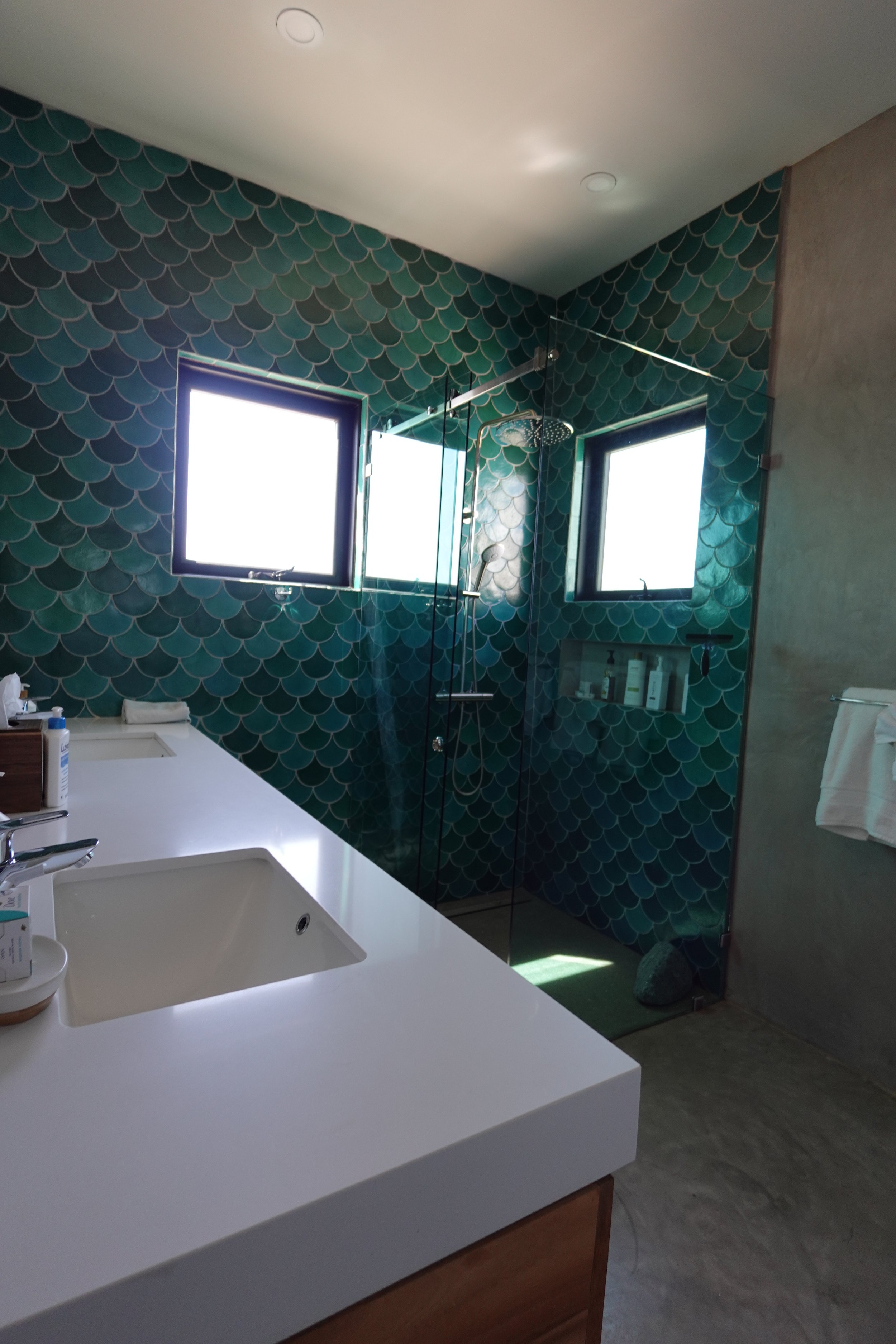 Modern bathroom with teal fish-scale patterned tiles, two square windows, a glass shower enclosure with a rainfall showerhead, a white countertop with a sink, and a towel hanging on a rack.