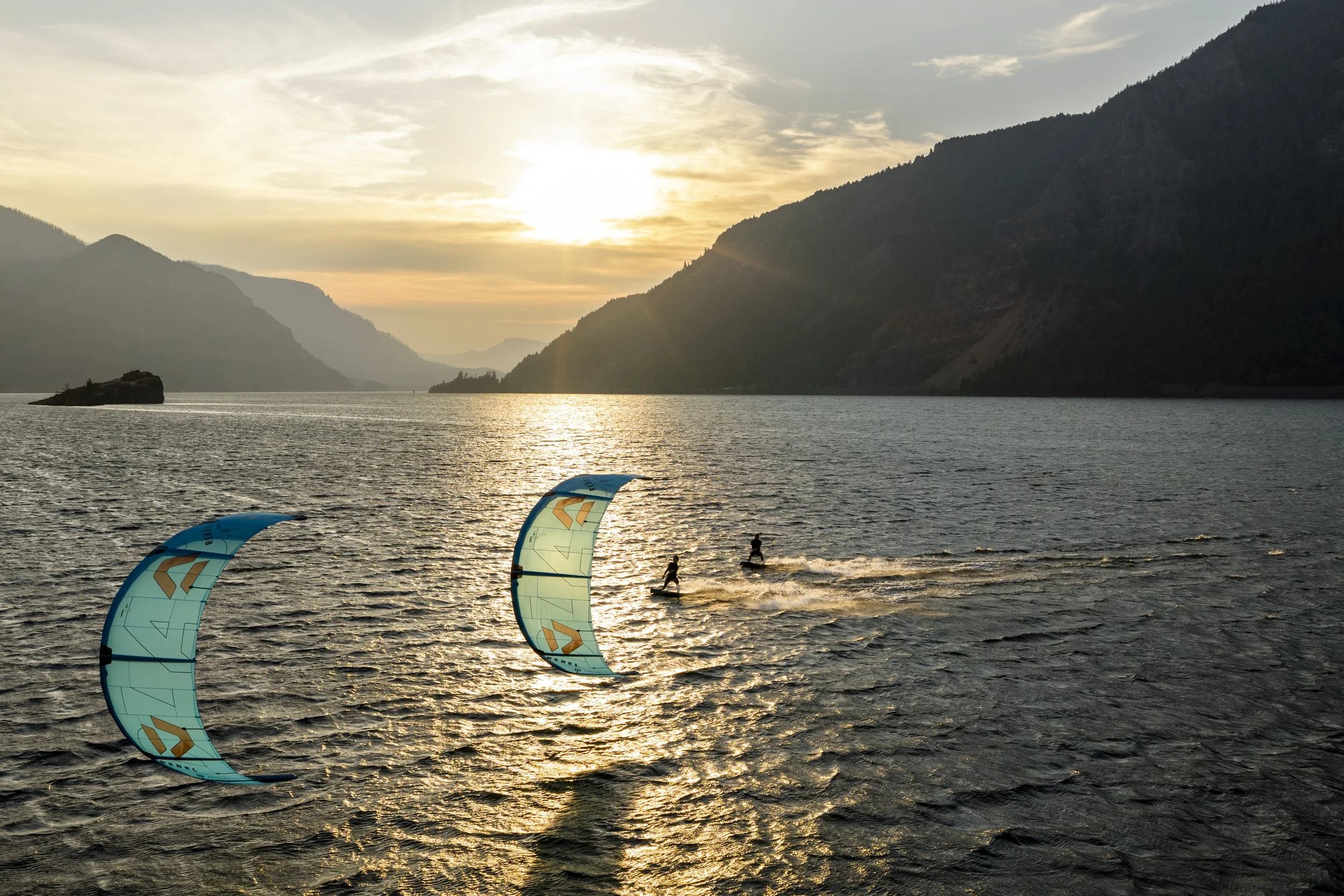 A sunset over a body of water with mountains in the background, and three kite surfers riding the waves with their blue and orange kite sails in the foreground.
