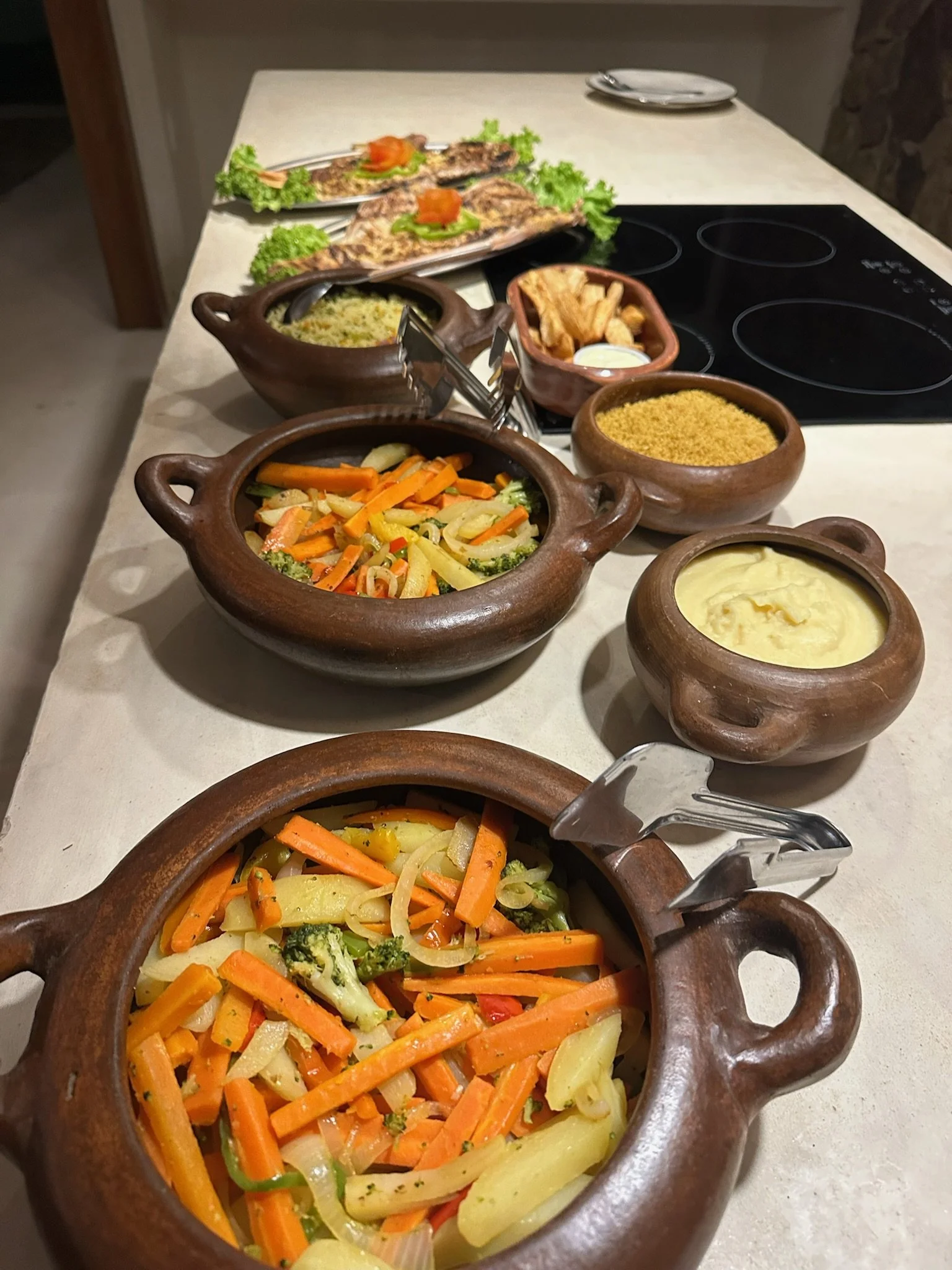 Baked vegetables and rice served in brown clay pots, with grilled fish garnished with lettuce and tomato slices in the background on a white countertop.