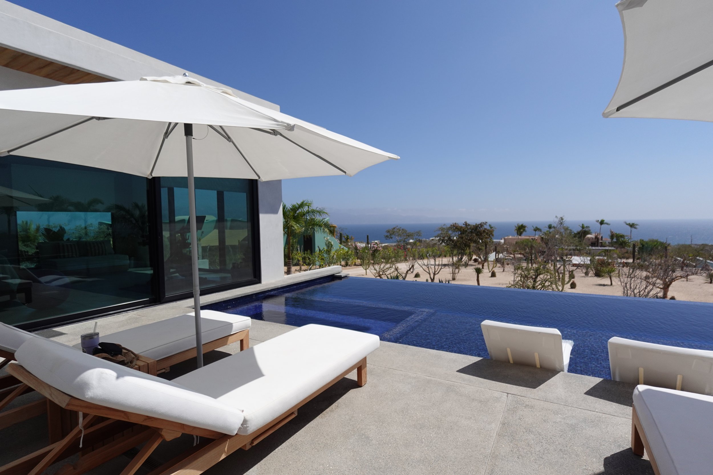Modern outdoor pool area with white lounge chairs under large white umbrellas, overlooking a desert landscape with sparse trees, houses, and the ocean in the distance under a clear blue sky.