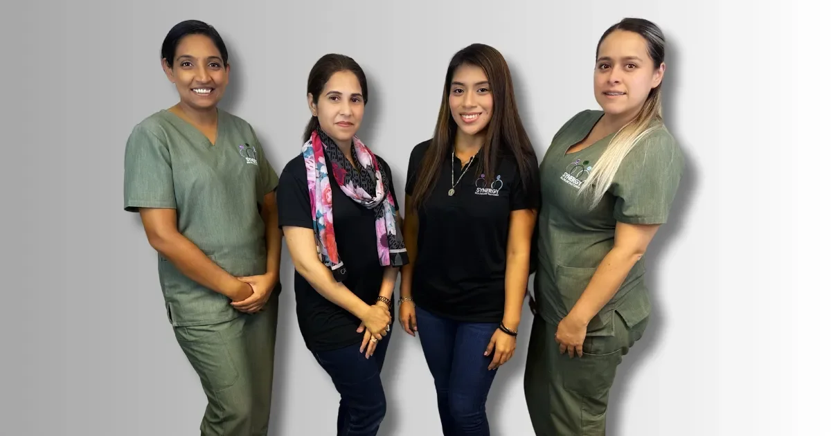 Cuatro mujeres de pie junto a una pared gris, dos con uniformes verdes, una con blusa negra y bufanda colorida, y otra con camiseta negra y jeans.