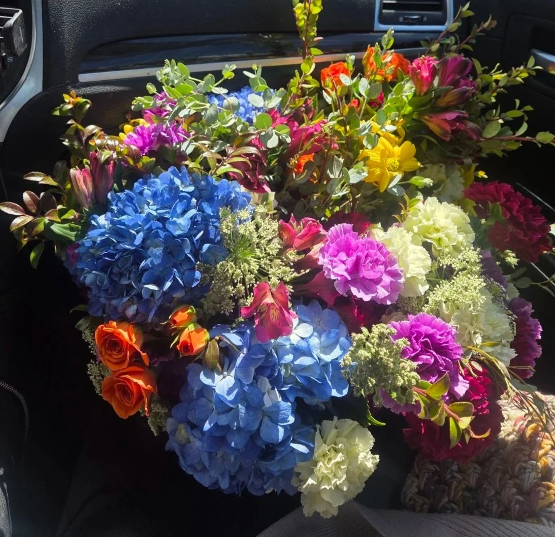 Large colorful bouquet of flowers, including blue hydrangeas, pink carnations, orange roses, yellow daisies, and other mixed blooms, placed on a car seat.