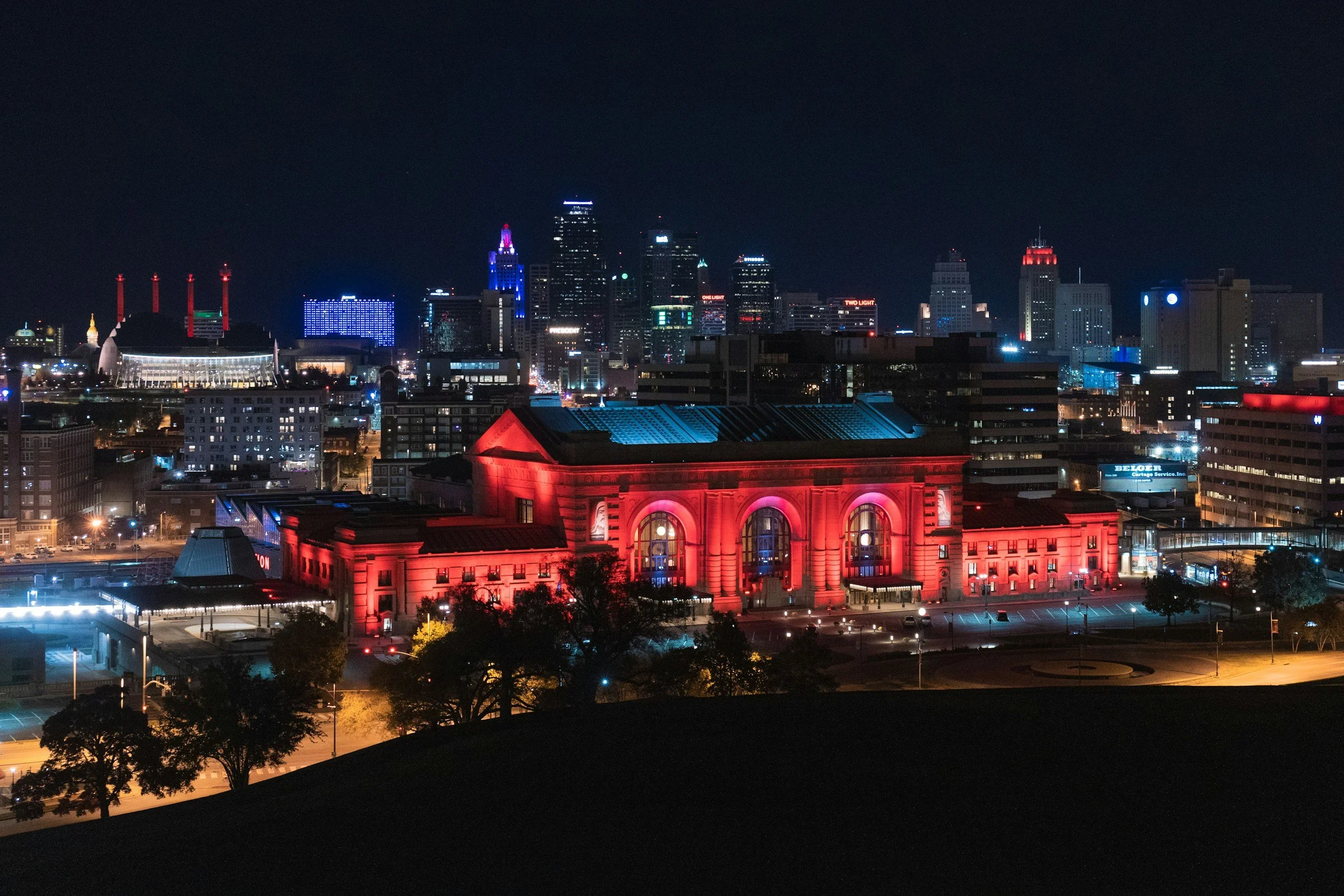 Nighttime cityscape featuring a large, historic building illuminated in red in the foreground, with a modern skyline in the background, including various skyscrapers and buildings with colorful lights.