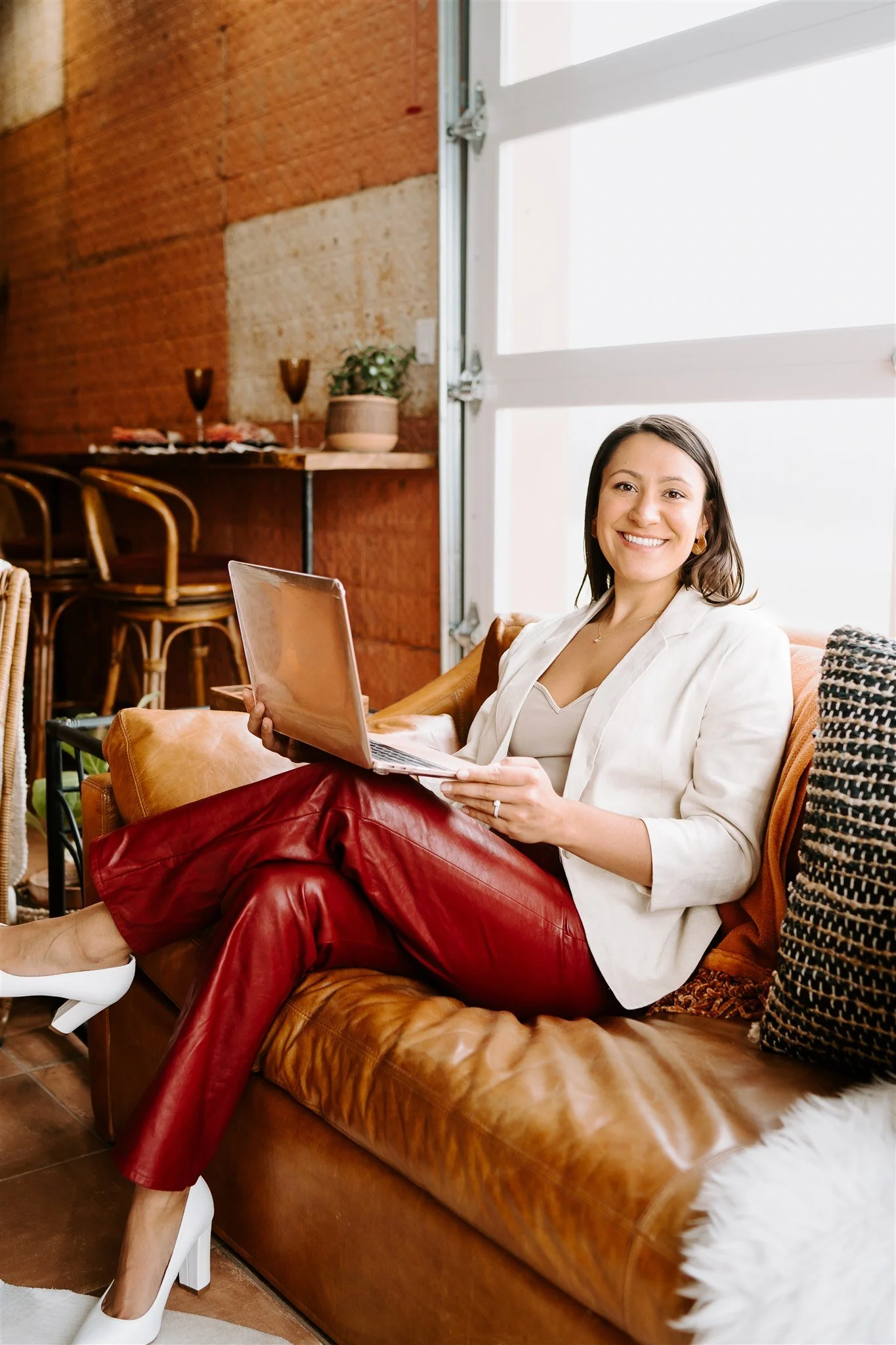 A woman sitting on a brown leather couch with a laptop, smiling at the camera. She is dressed in a white blazer, red leather pants, and high heels. There is a window behind her, and the room has a rustic interior with exposed brick walls and decorative items.