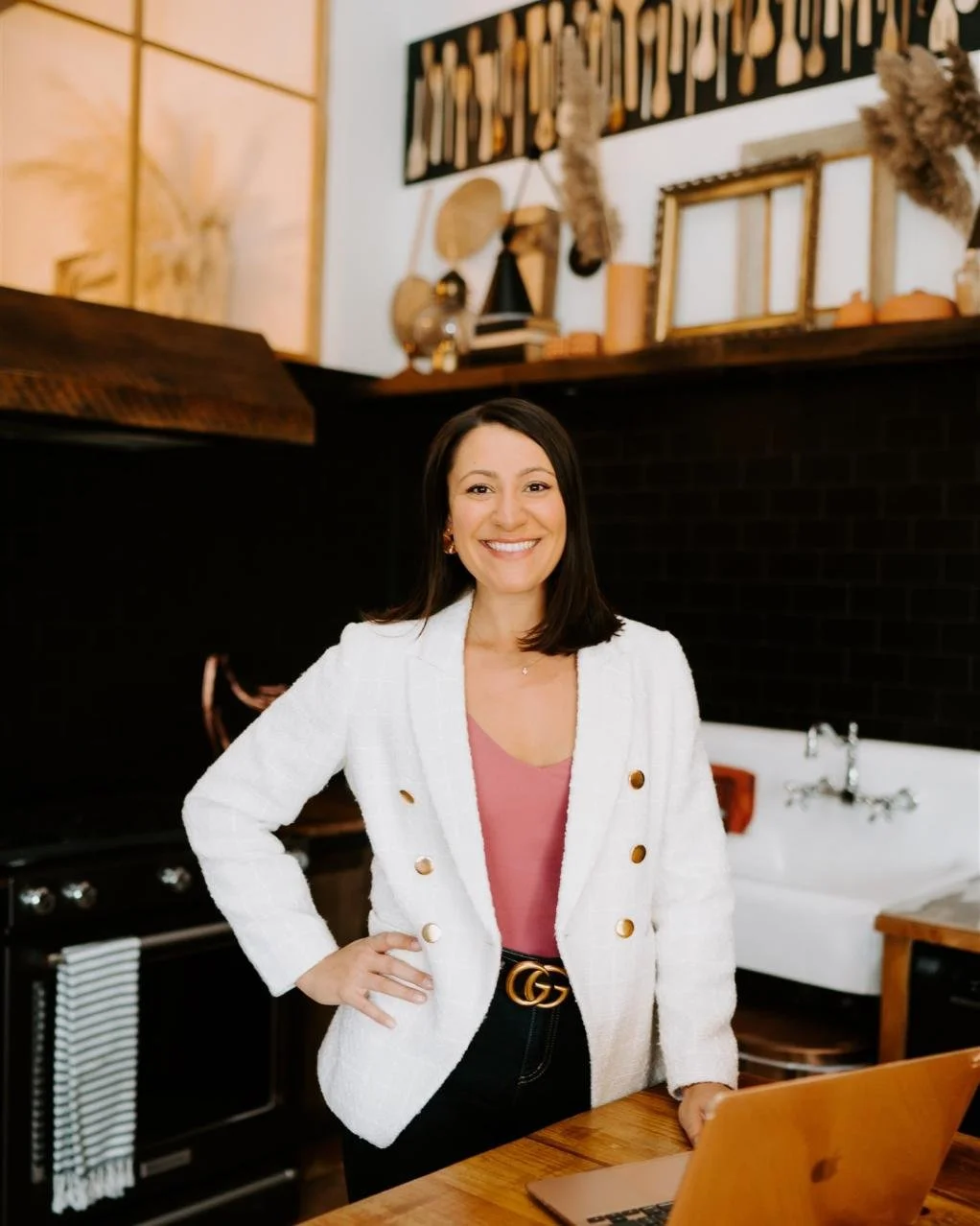 A woman with shoulder-length dark hair smiling while standing in a kitchen, wearing a white blazer with gold buttons, a pink top, and black pants with a Gucci belt. There is a laptop on a wooden table in front of her, and the background features decorative wall art, dried plants, and a kitchen sink.