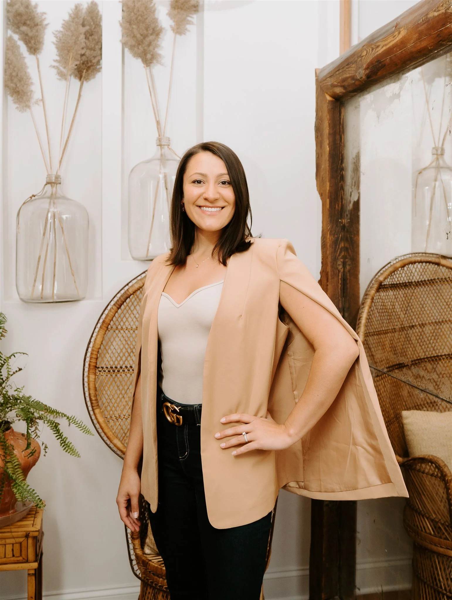 A woman standing indoors with a smile, wearing a beige blazer over a white top, black pants, and a belt with a gold buckle, with wavy brown hair.