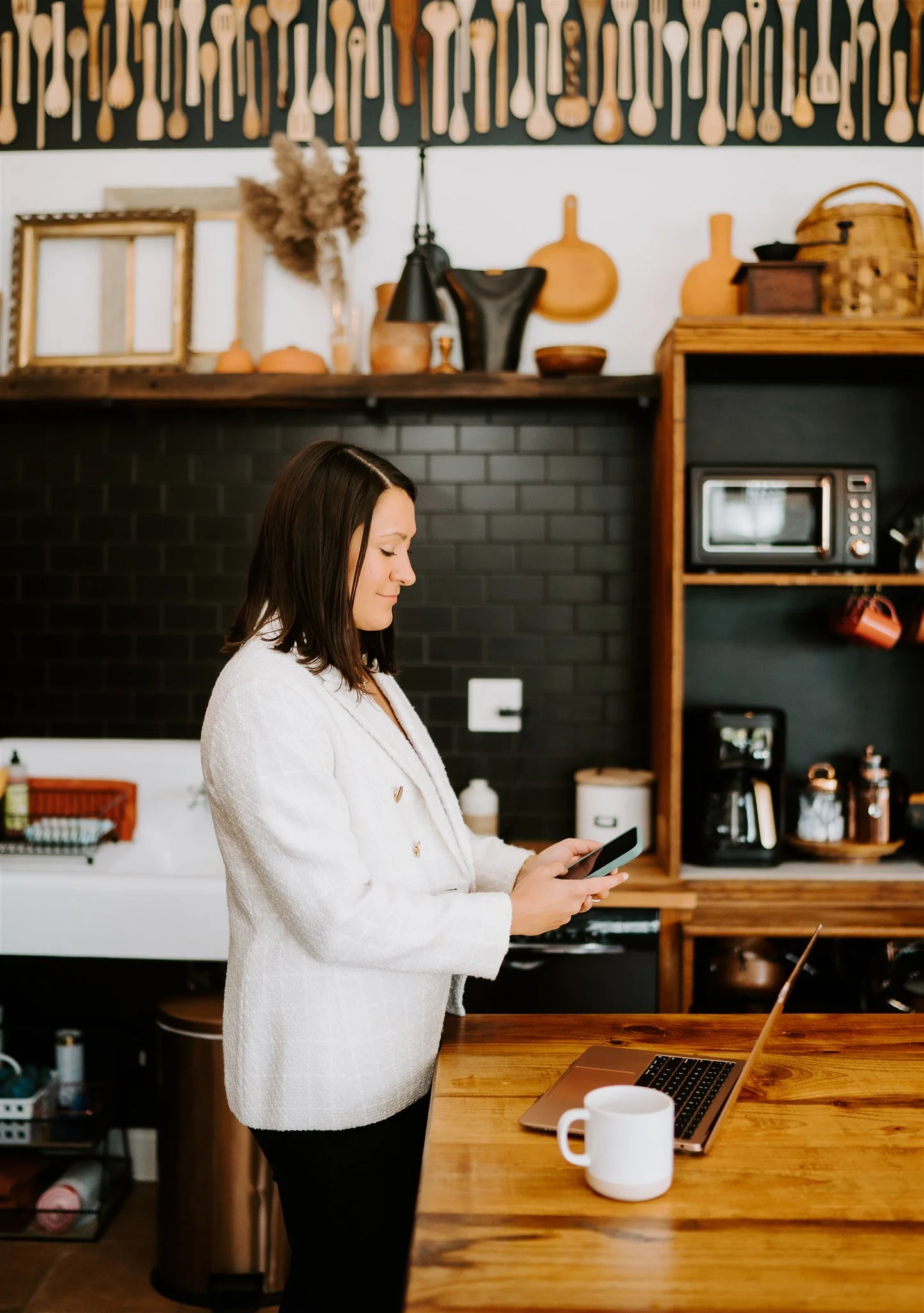 A woman with shoulder-length dark hair stands in a modern kitchen, looking at her smartphone. The kitchen has a black brick wall, wooden shelves with kitchen items, a microwave, coffee maker, and a laptop on the wooden counter. A white mug is on the counter, and a laptop with a brown cover is open. The background features decorative wooden utensils and framed pictures.