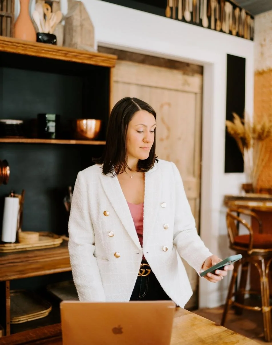 A woman in a white blazer is standing in a cozy kitchen, looking at her phone, with a laptop on the table in front of her.