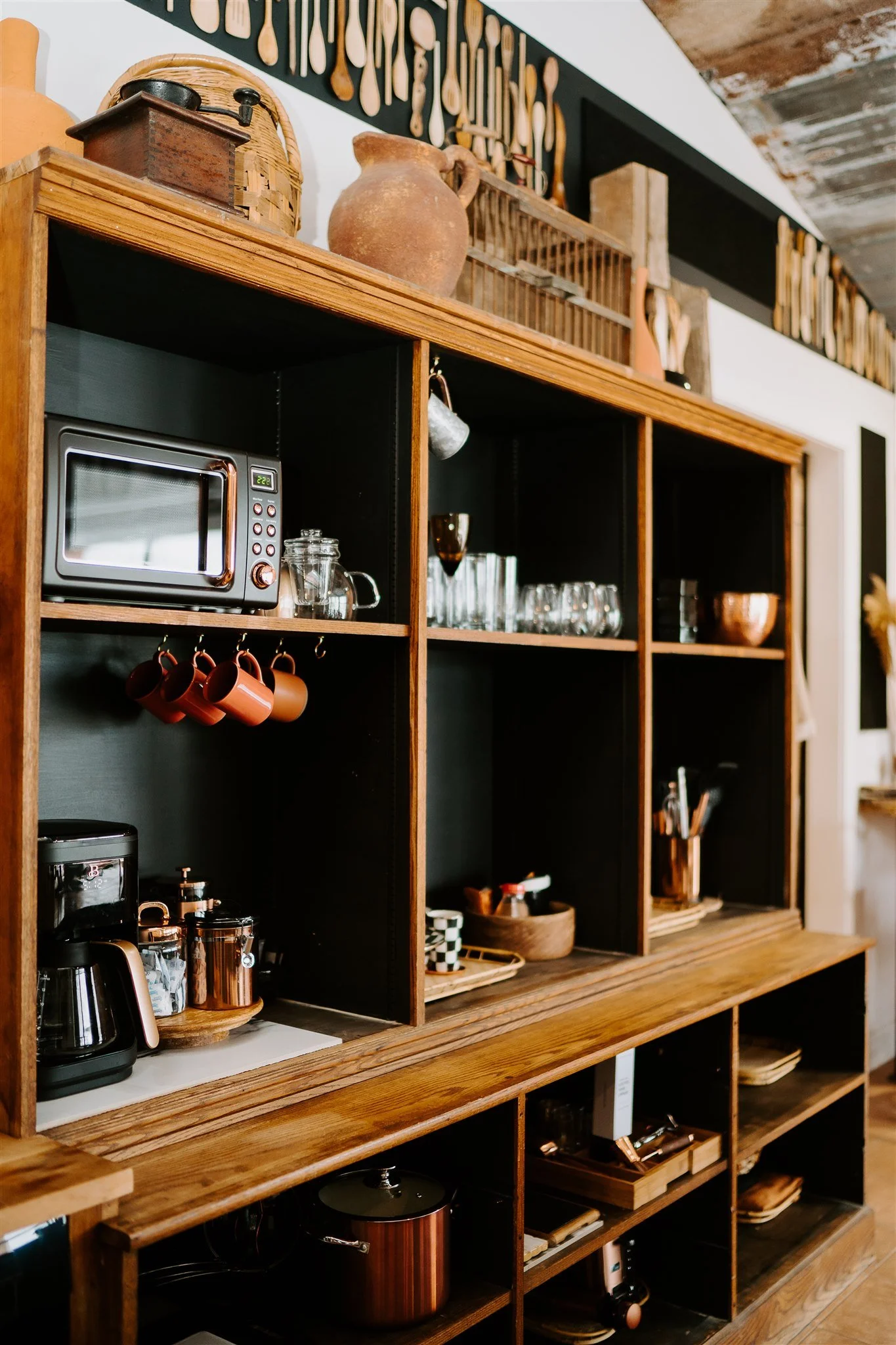 Wooden kitchen shelving unit with various kitchen items, including a microwave, glassware, copper pots, and cups, with decorative items on top and utensils hanging on the wall above.