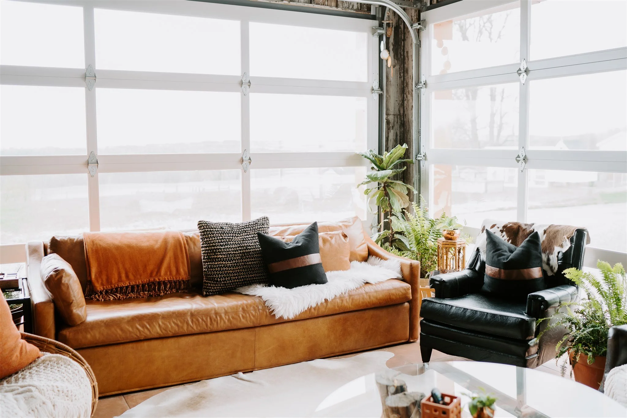 Living room with large front windows, tan leather sofa with pillows, black armchair, various plants, and cozy decor.