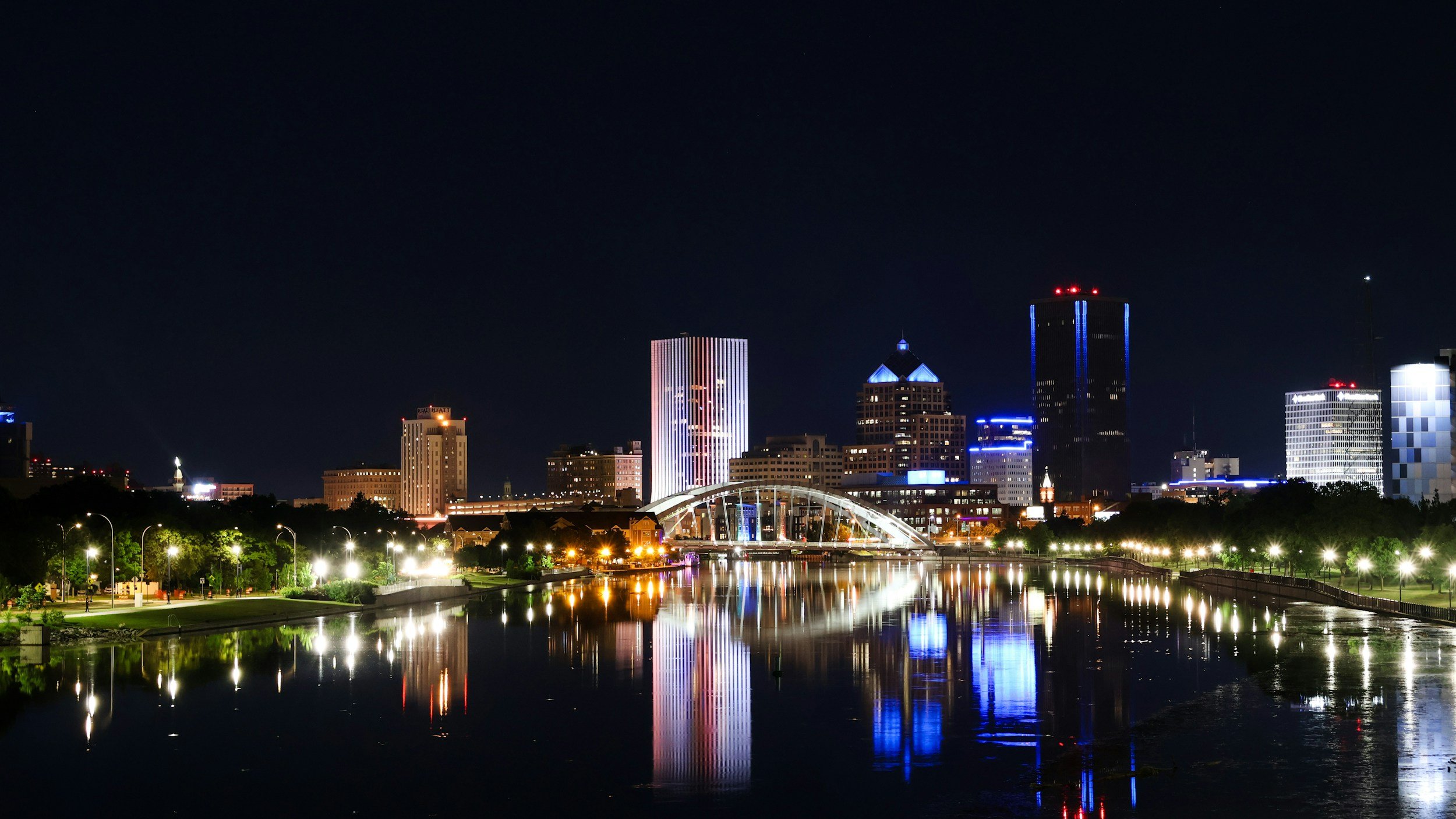 Nighttime city skyline with illuminated skyscrapers and their reflections on a river.