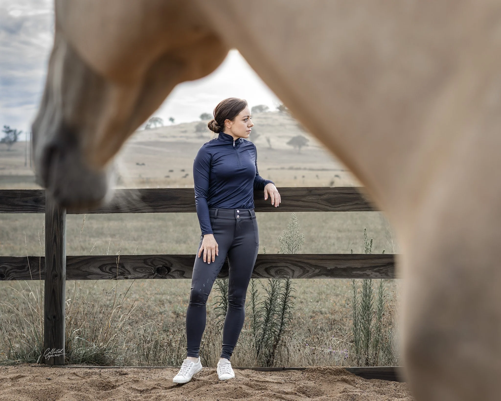 This picture shows a woman in focus leaning against a fence, above her and slightly out of focus, we see the underneath of a horses head and neck, framing the shot.