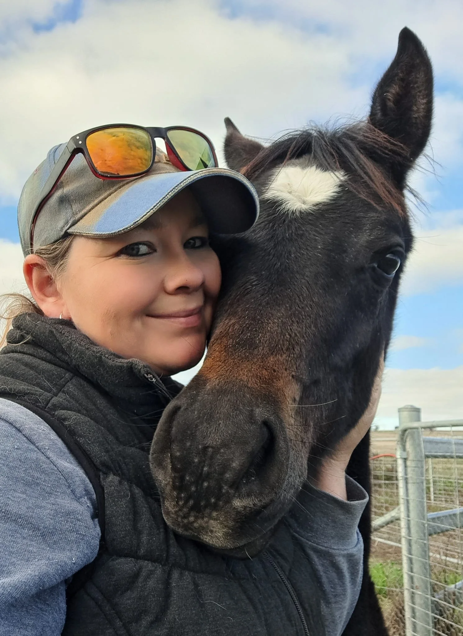 The image shows a girl with a cap and sunglasses on, cuddling the face of a foal who is also looking at the camera