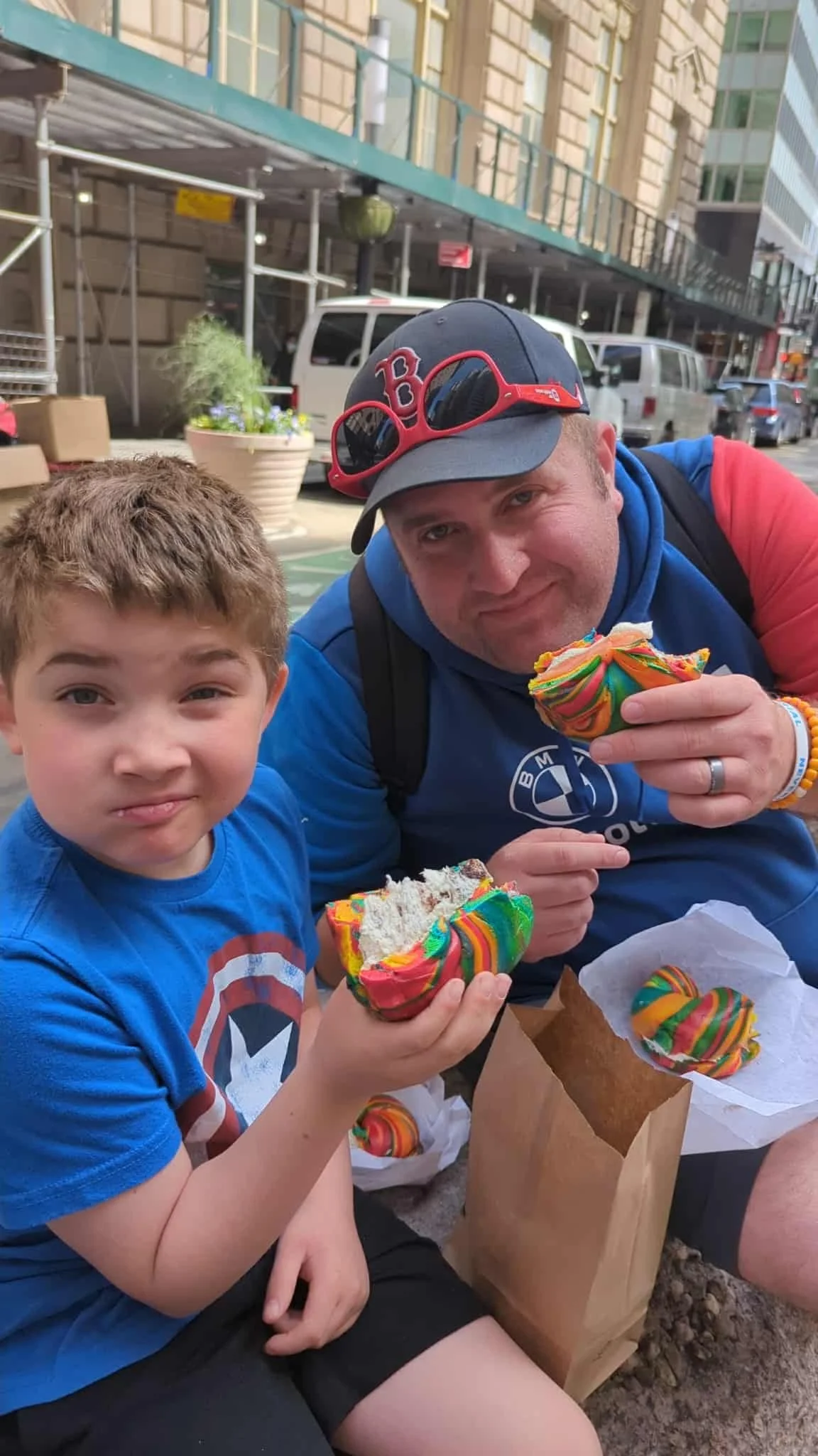 Mark and his son, sitting on the ground outdoors, eating colorful rainbow pastries.