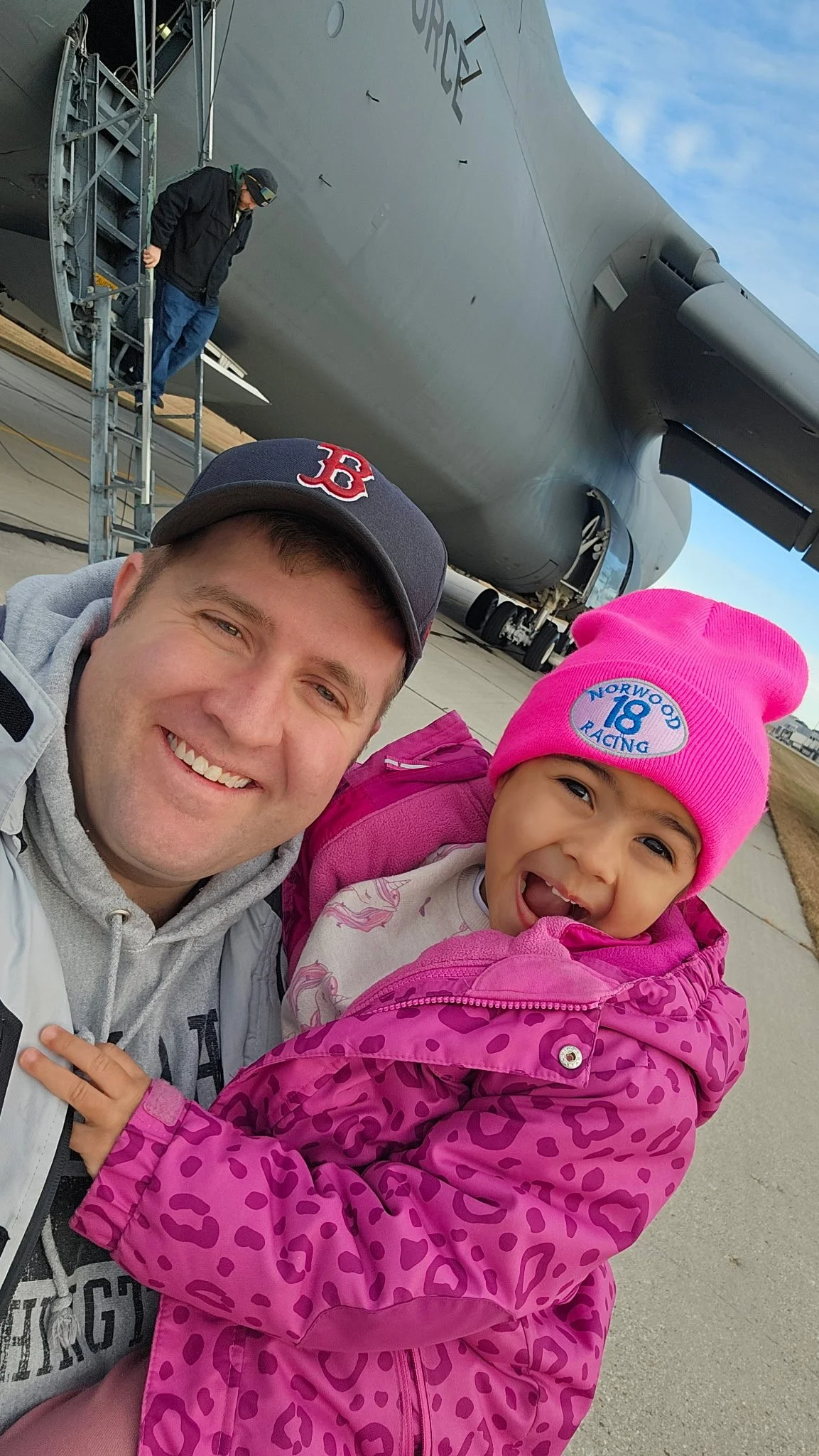 Mark and his daughter taking a selfie in front of a large military airplane on an airfield. Mark is wearing a navy Boston Red Sox cap and a gray jacket; his daughter is in a pink leopard-print jacket and pink beanie.