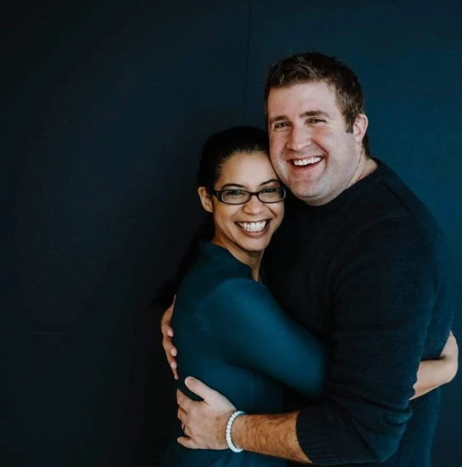 Mark and his wife hugging each other against a dark blue background.