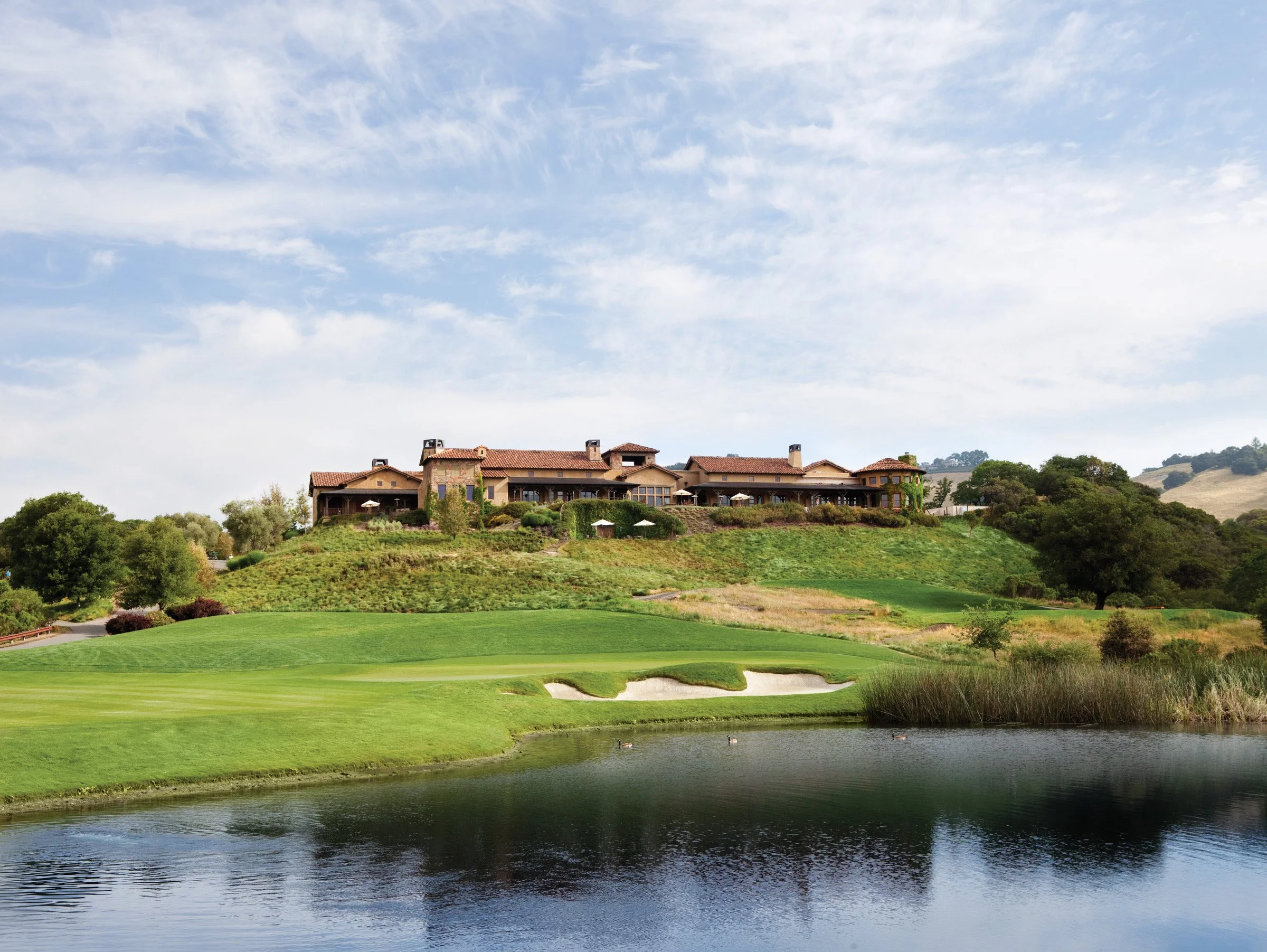 A golf course with a pond in the foreground, lush green grass, and a large, multi-story building with a red-tiled roof on a hill in the background under a partly cloudy sky.