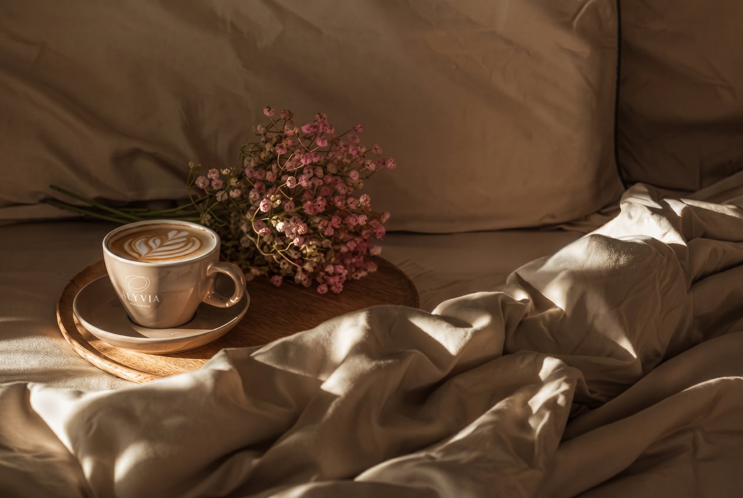 A hotel room with a beige tufted armchair, a bed with white linens and a pillow, a wooden nightstand with a telephone, a wall-mounted reading lamp, and a modern round yellow hanging lamp against a wooden-paneled wall. There are sheer curtains hiding a window.