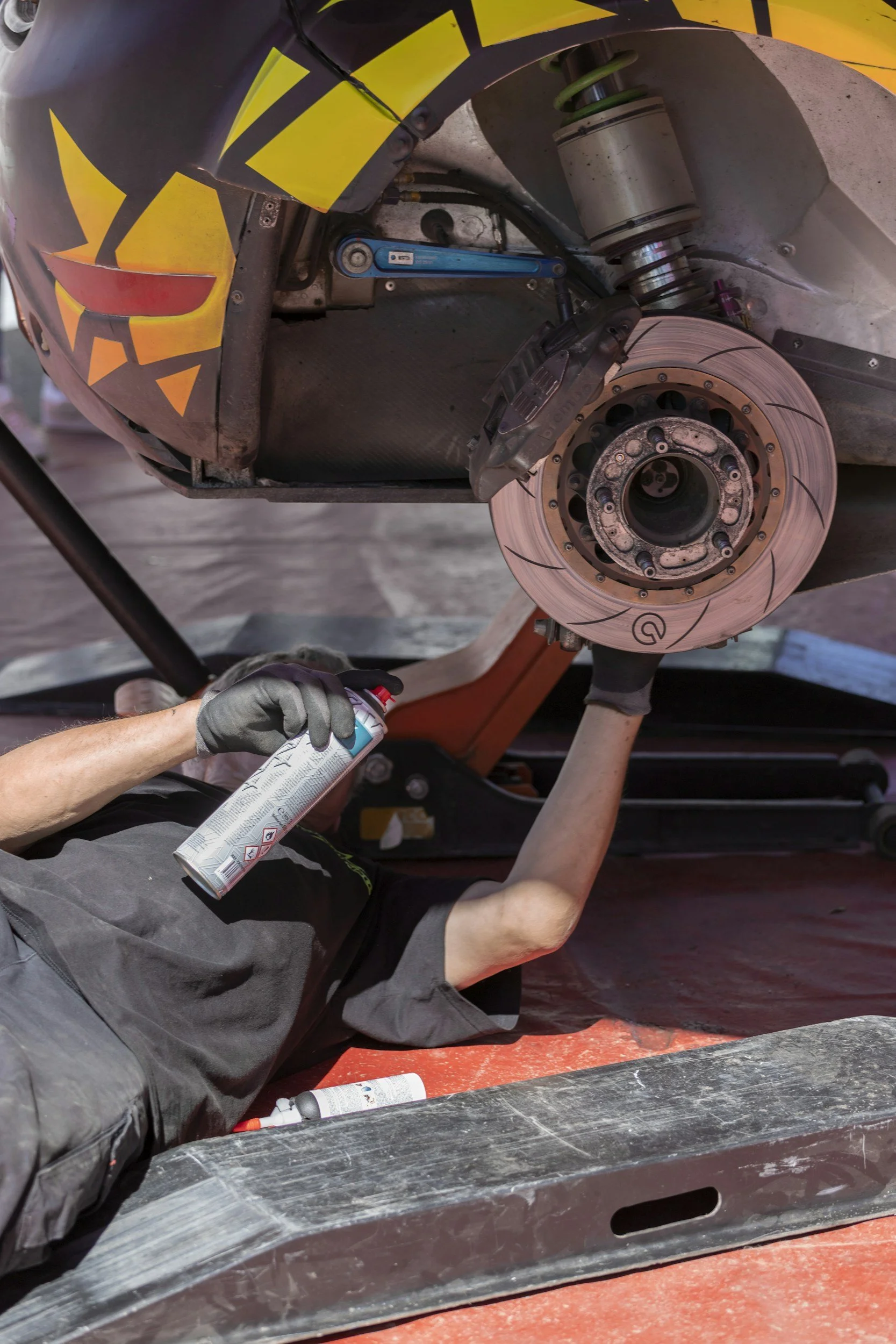 A person lying on the ground working on a race car, holding a spray can, with a visible suspension and brake system.