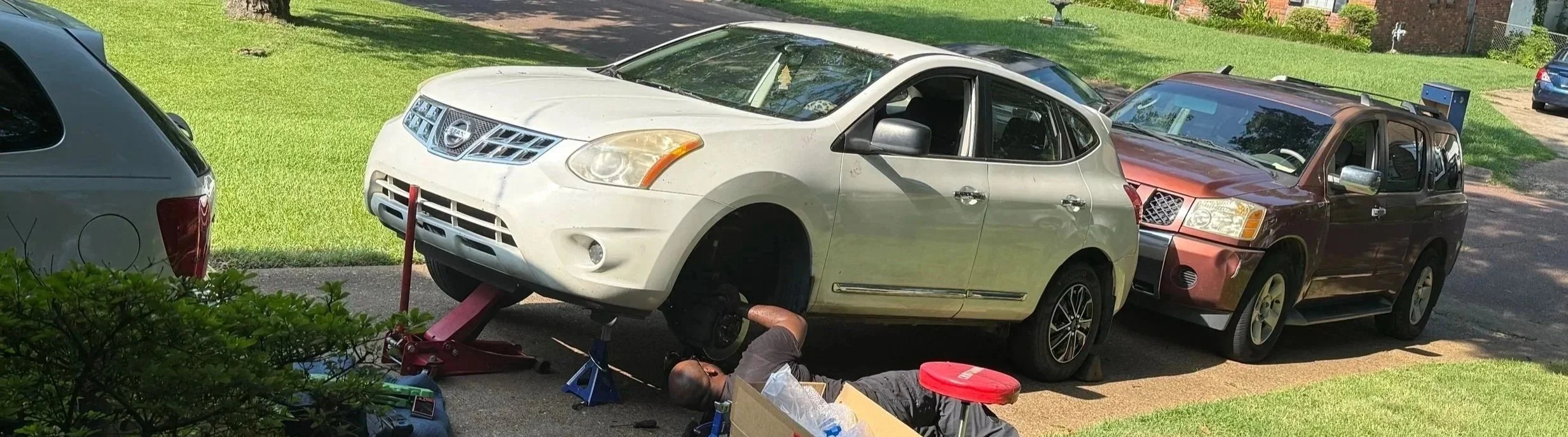 A person is working underneath a white Nissan hatchback car, which is elevated with a red car jack. The car's front wheel is removed. There are two other parked vehicles nearby on a grassy area, with shadows indicating a sunny day.