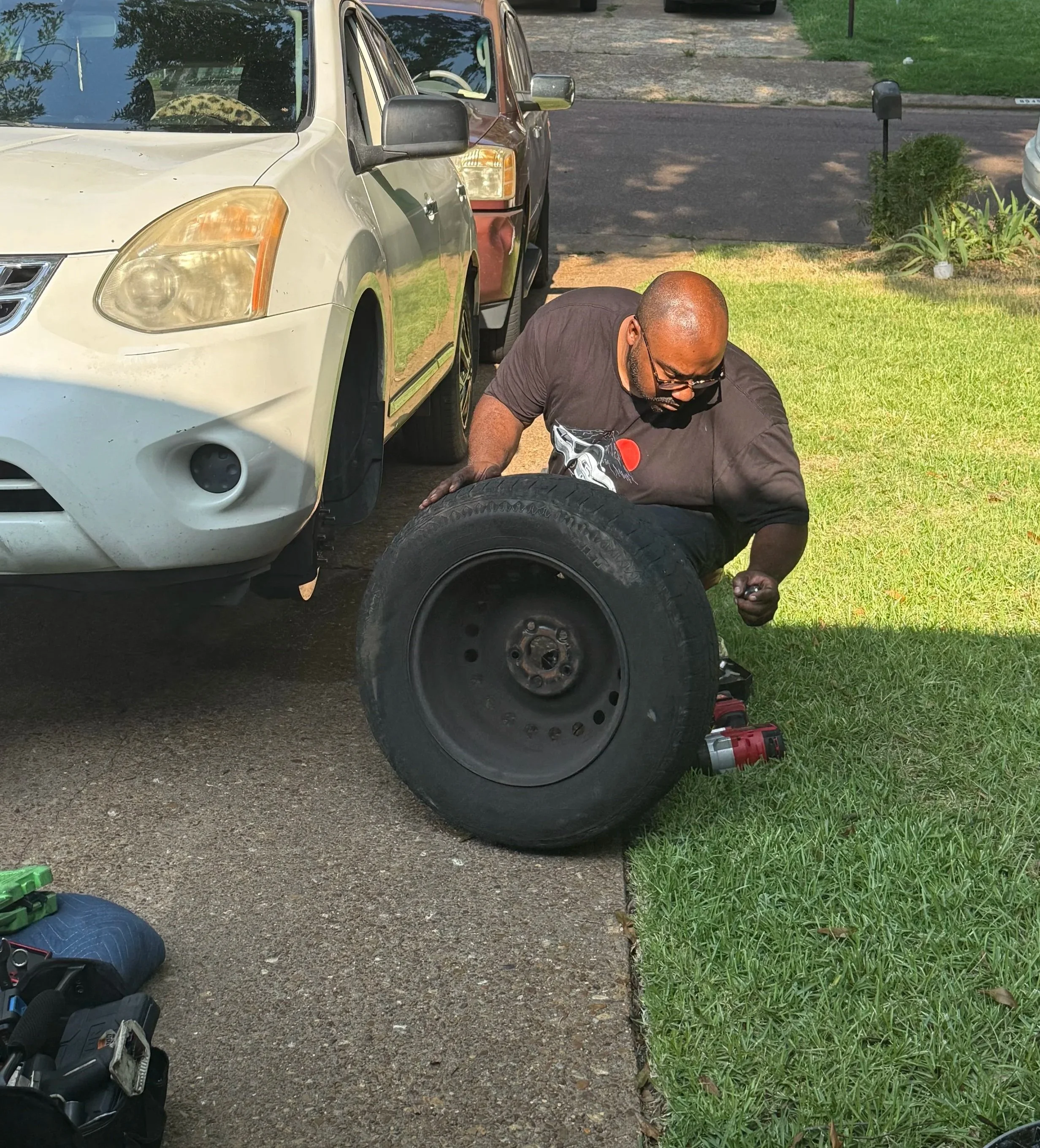 A man fixing a flat tire on a car parked on a driveway, with tools and a green toolbox nearby, in a residential area with grass and a sidewalk.